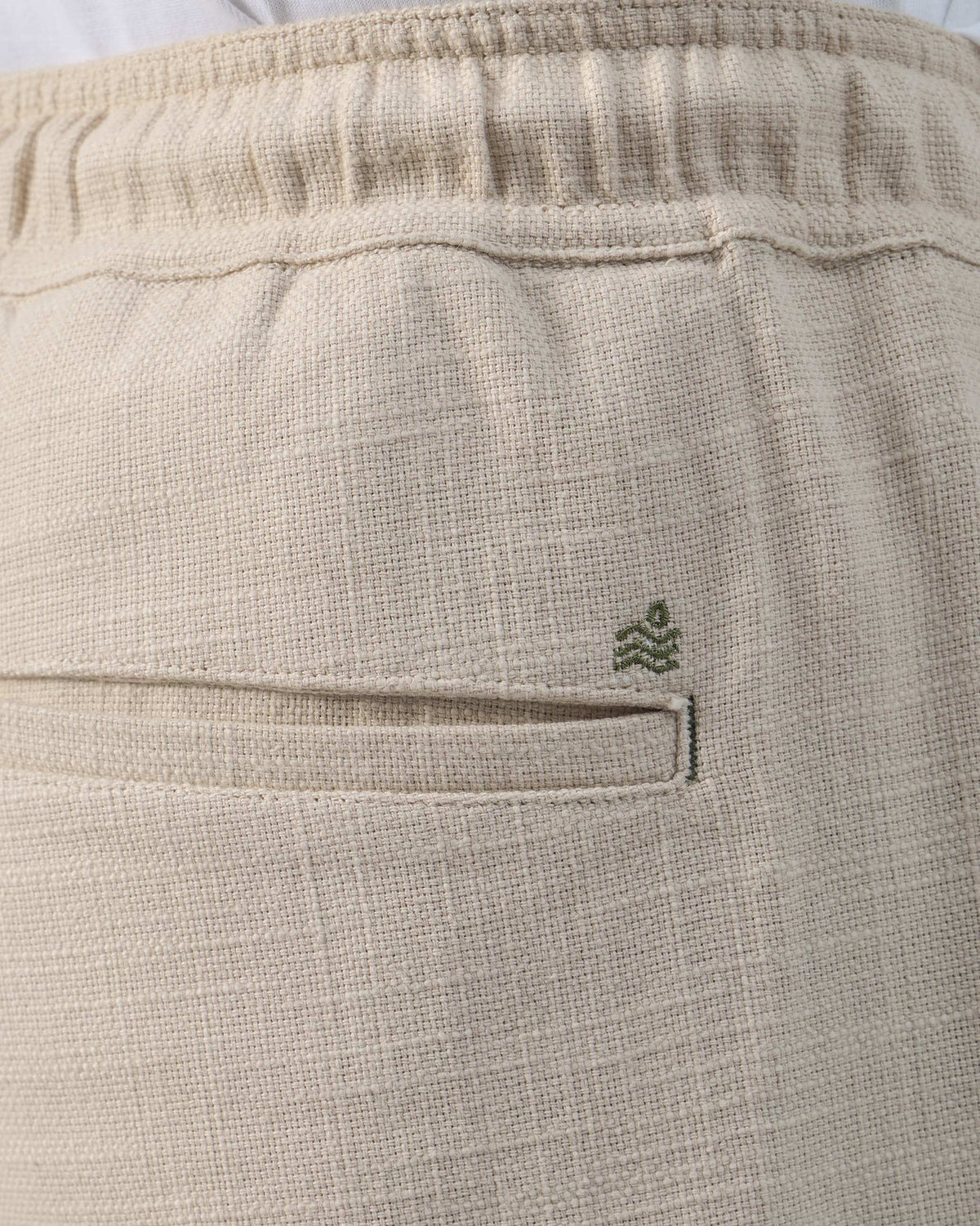 Close-up of a beige Breeze trouser welt pocket with a green leaf logo.