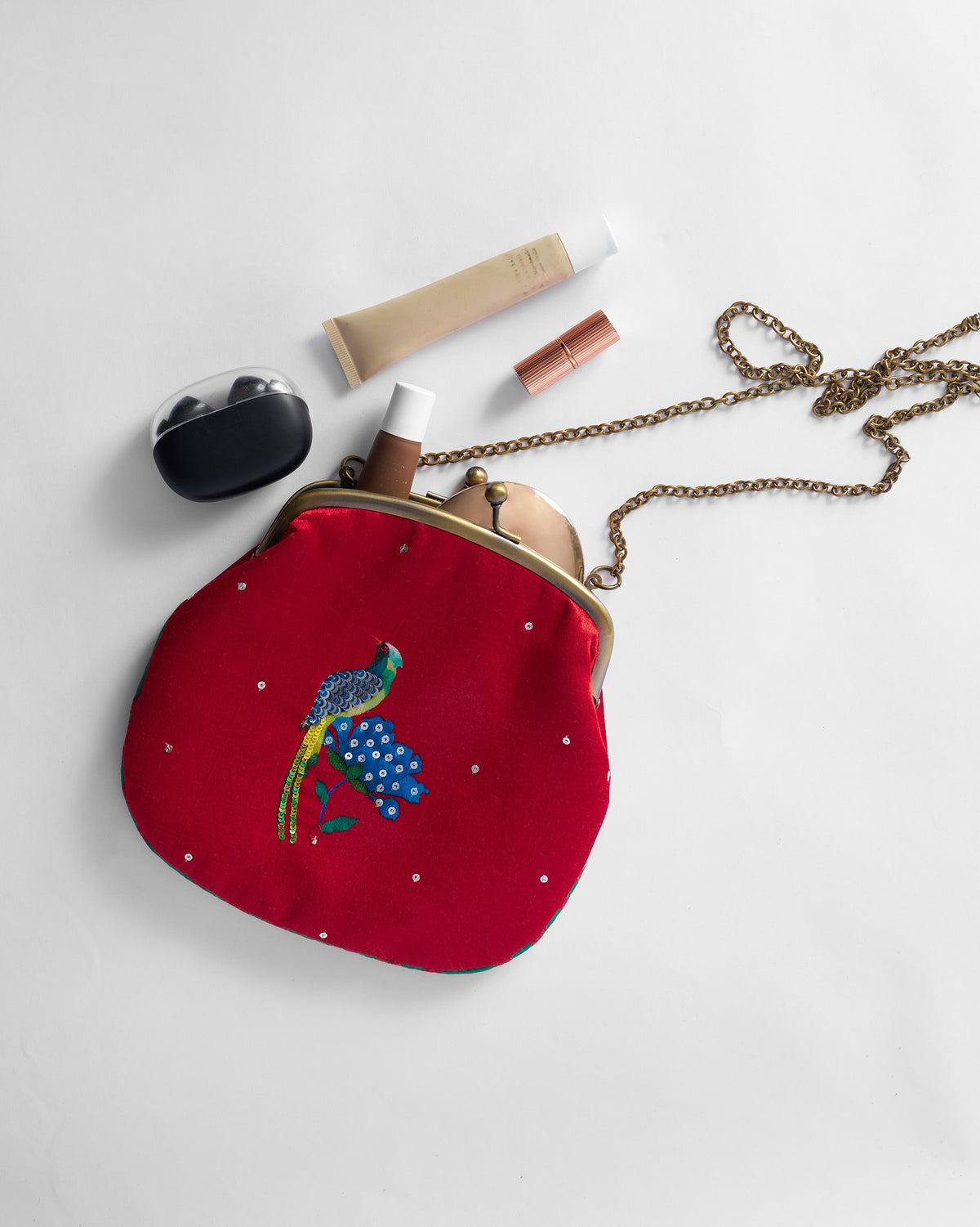 Red bird-embroidered clutch open beside lipstick, compact, and earbuds.