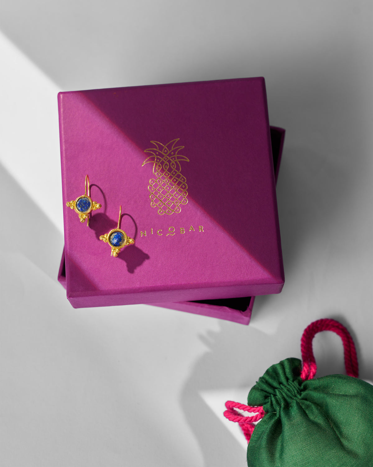 Pair of gold Lapis Drops on a pink gift box beside a green pouch.