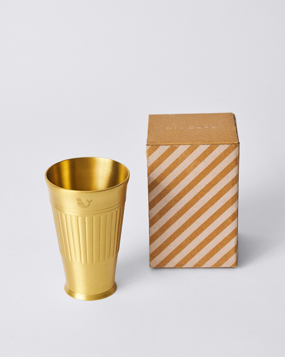 Brass Dariya lassi glass beside a striped Nicobar gift box.