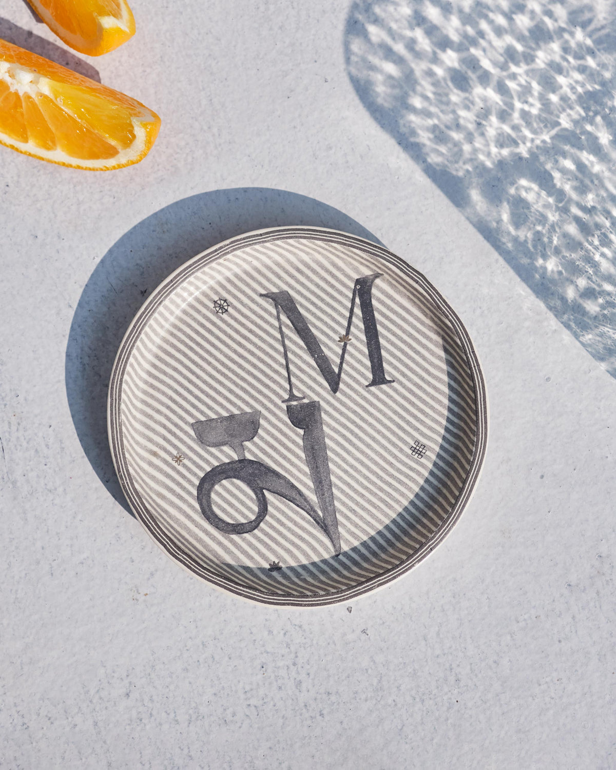 Cream Akshar coaster with a charcoal letter M and Devanagari म on diagonal stripes.