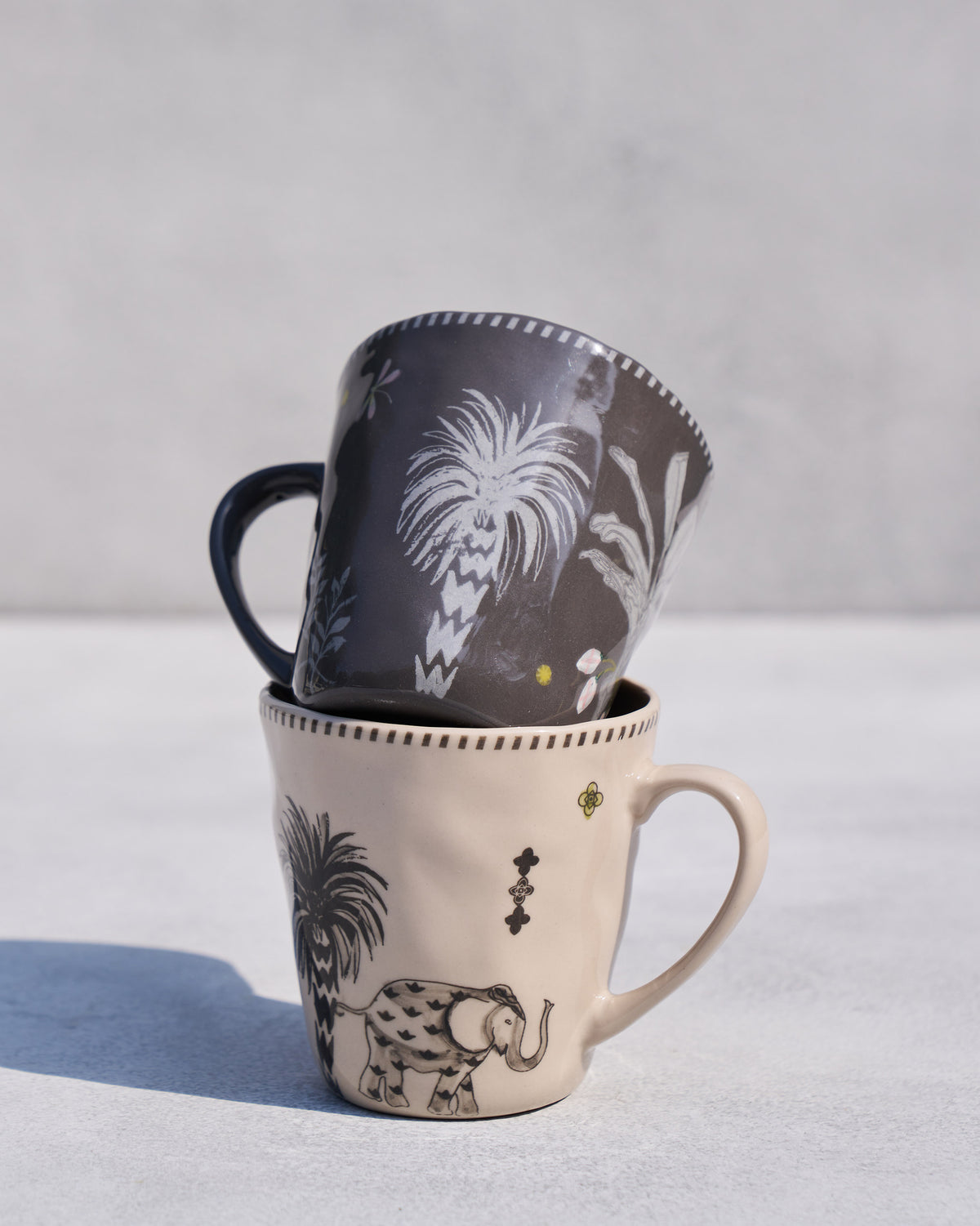 Stacked Malabar conical mugs showing charcoal palm and cream elephant designs.