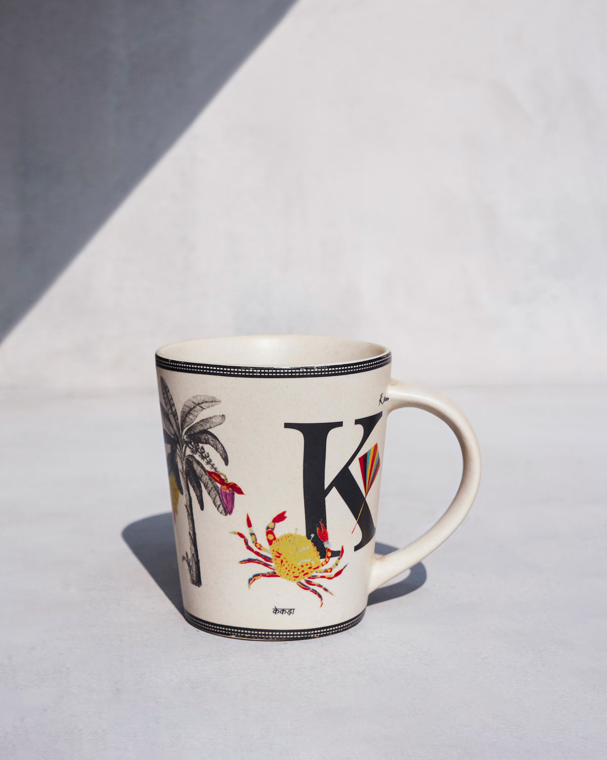 Cream Kamal mug with a black letter K, crab and kite motifs.