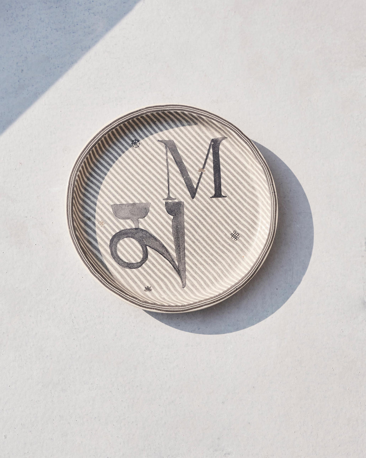 Cream Akshar coaster showing charcoal M and म on a fine striped ground.
