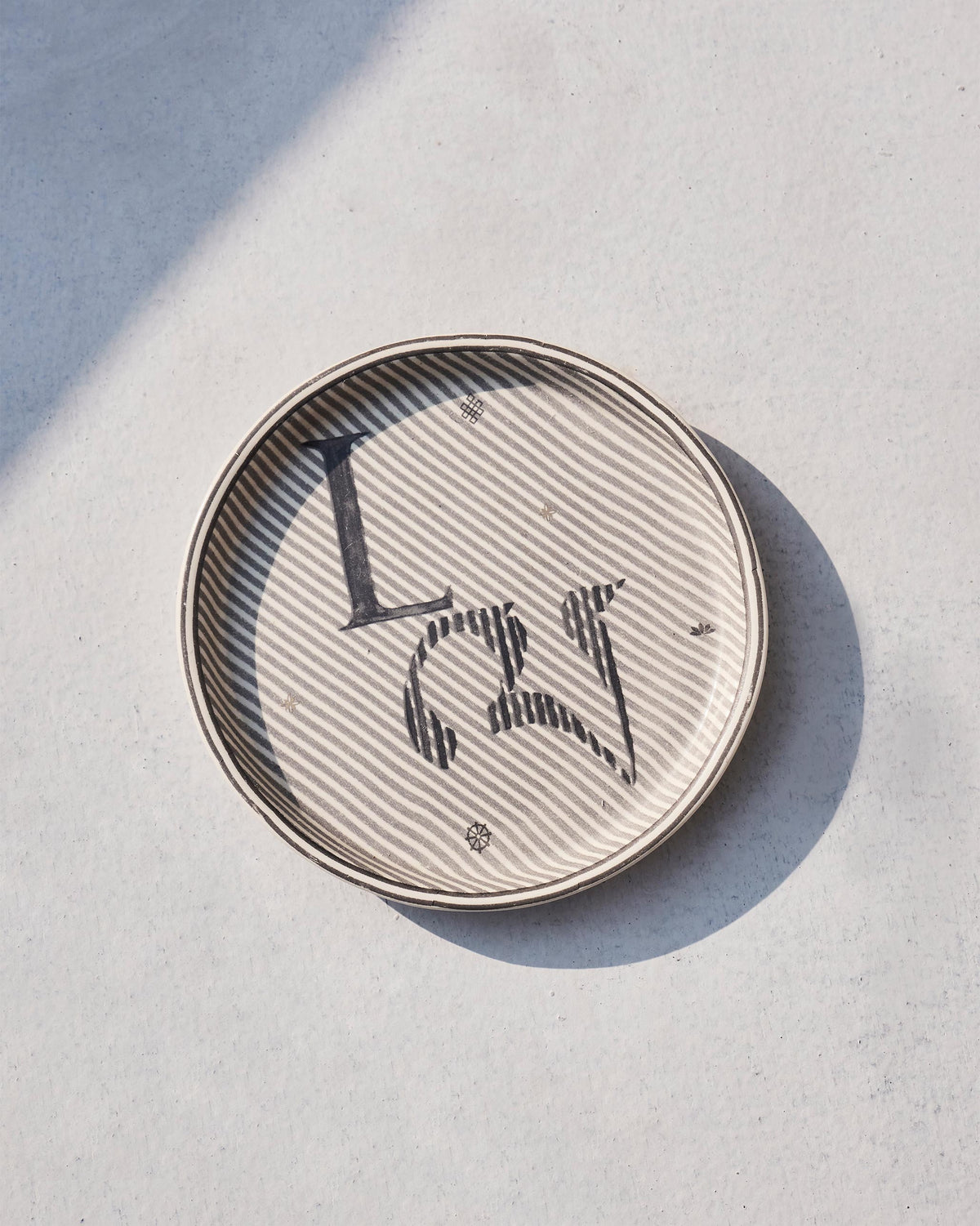 Akshar Coaster with a letter L on grey diagonal stripes, seen from above.