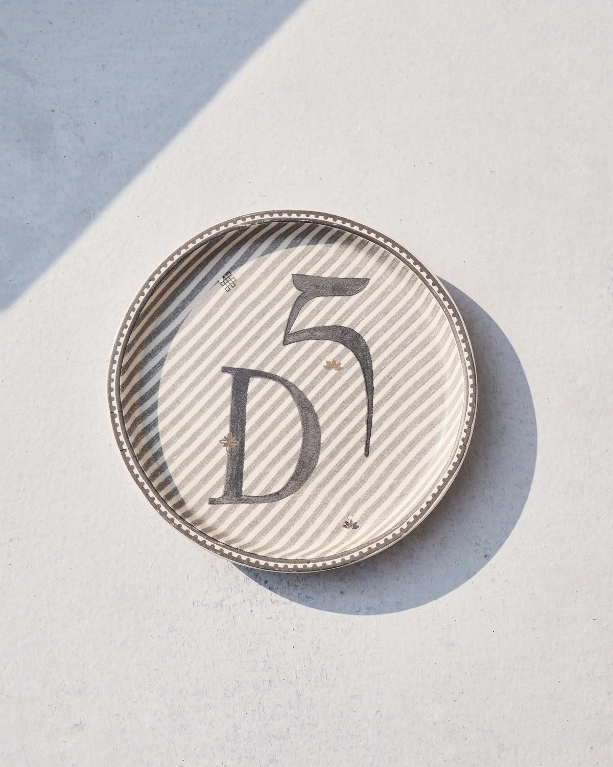 Akshar Coaster with a bold letter D on grey diagonal stripes, seen at an angle.