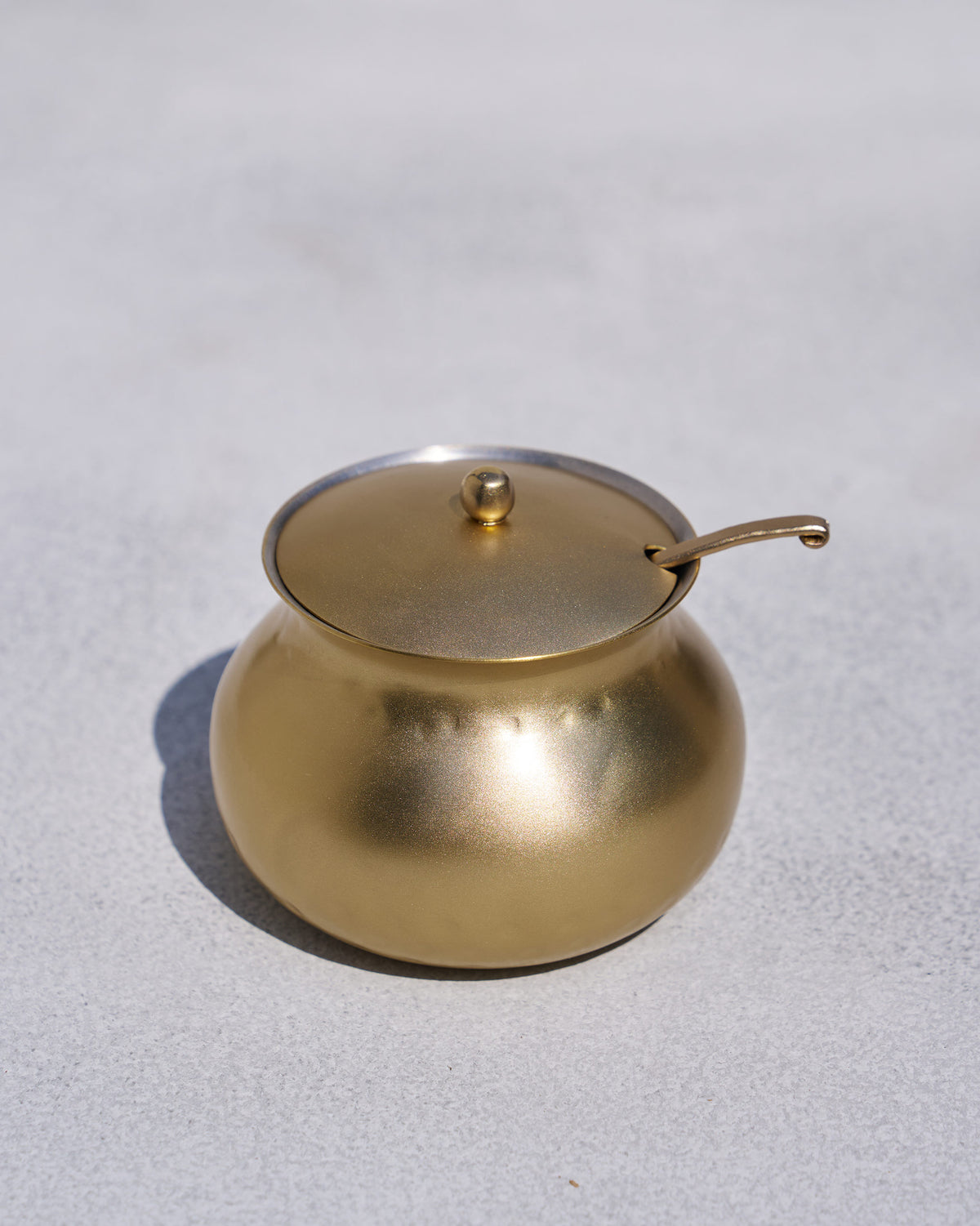 Rounded brass Madhu ghee pot with lid and ladle on a grey surface.