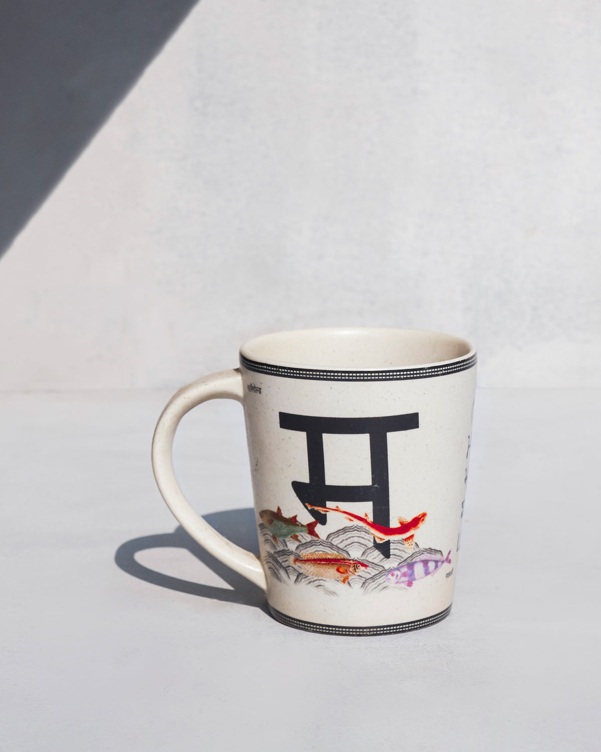 Cream Machli mug with a Devanagari letter and swimming fish.