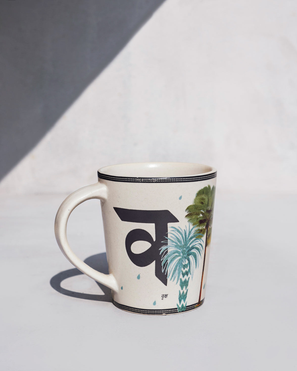 Cream Vriksh mug printed with Devanagari व and palm trees.