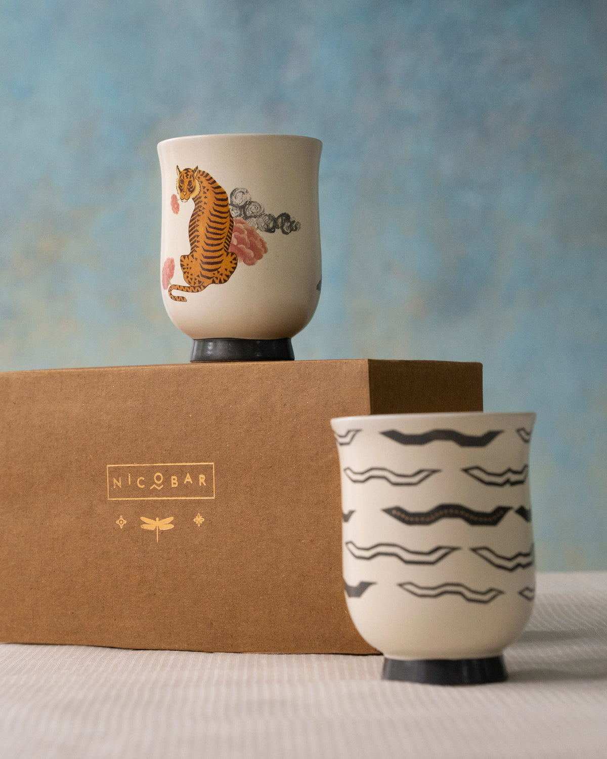 Sitara tea cups with a tiger and black waves, set beside a Nicobar box.