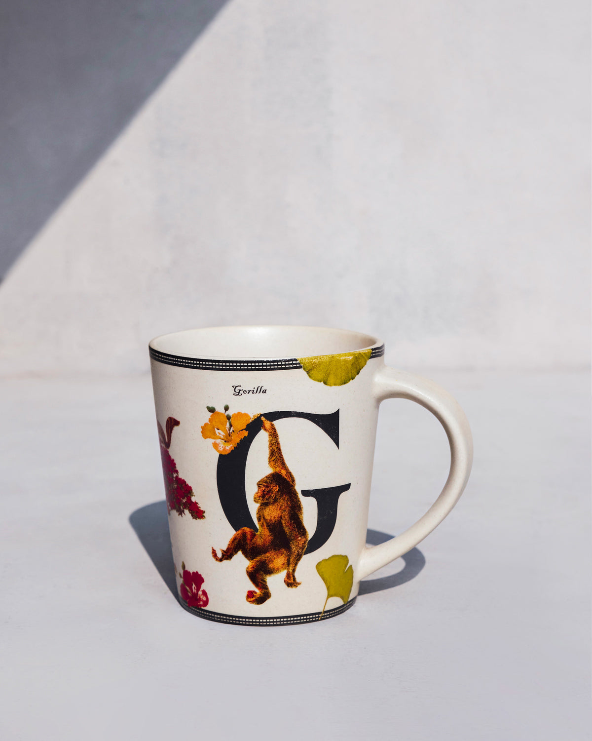 Cream Gulmohar mug with a black letter G and a gorilla among yellow flowers.