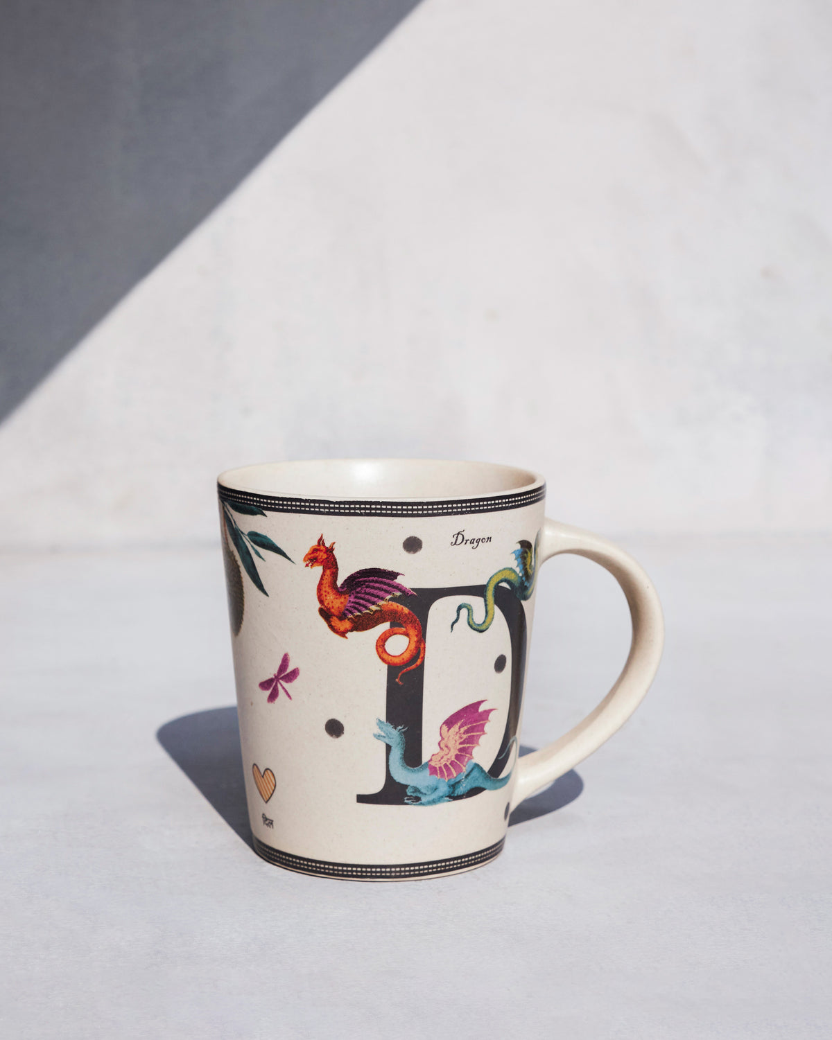 Cream Dariya mug with a letter D and colorful dragon illustrations.