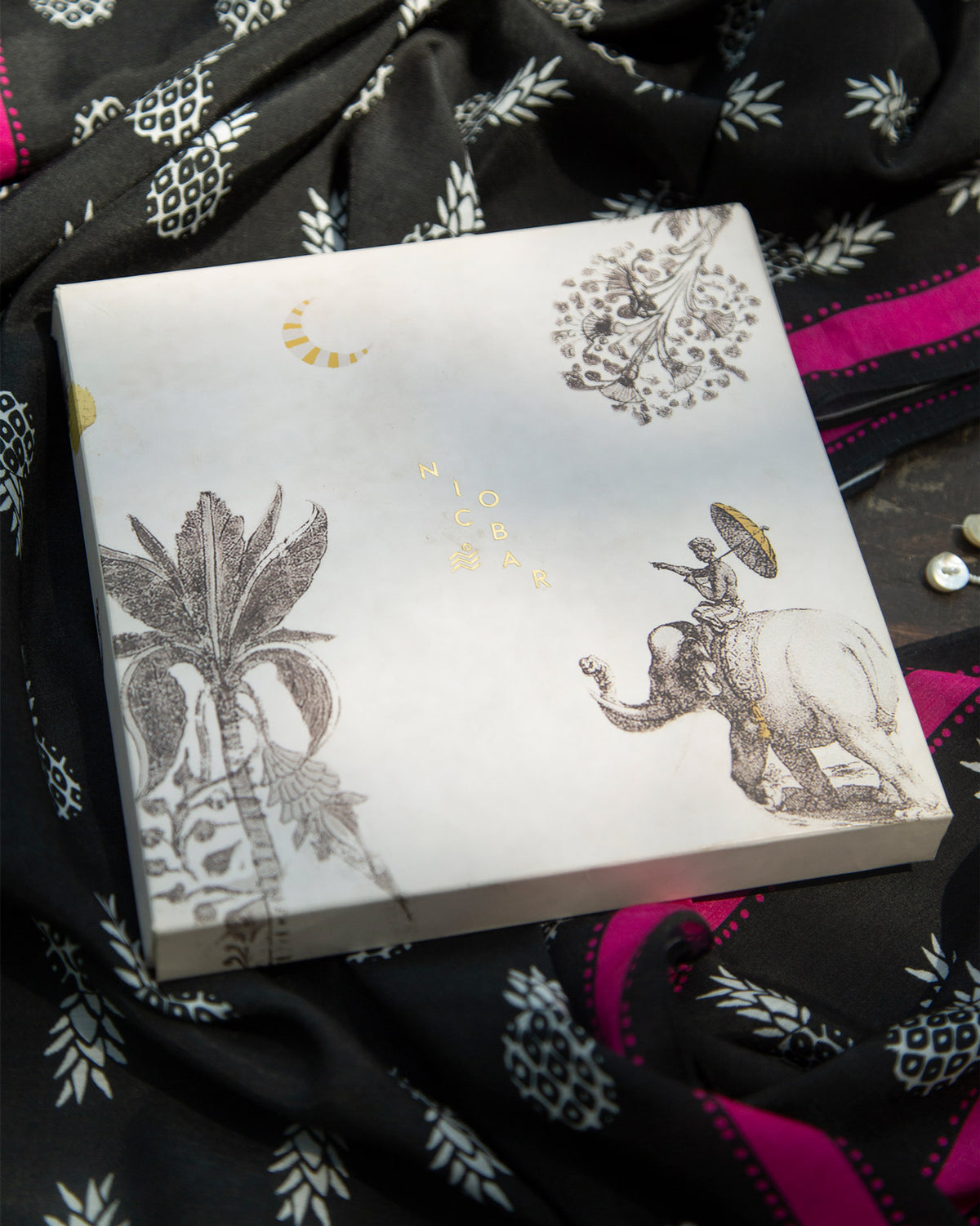 Nicobar gift box resting on a black Tropico Scarf with pink edges.