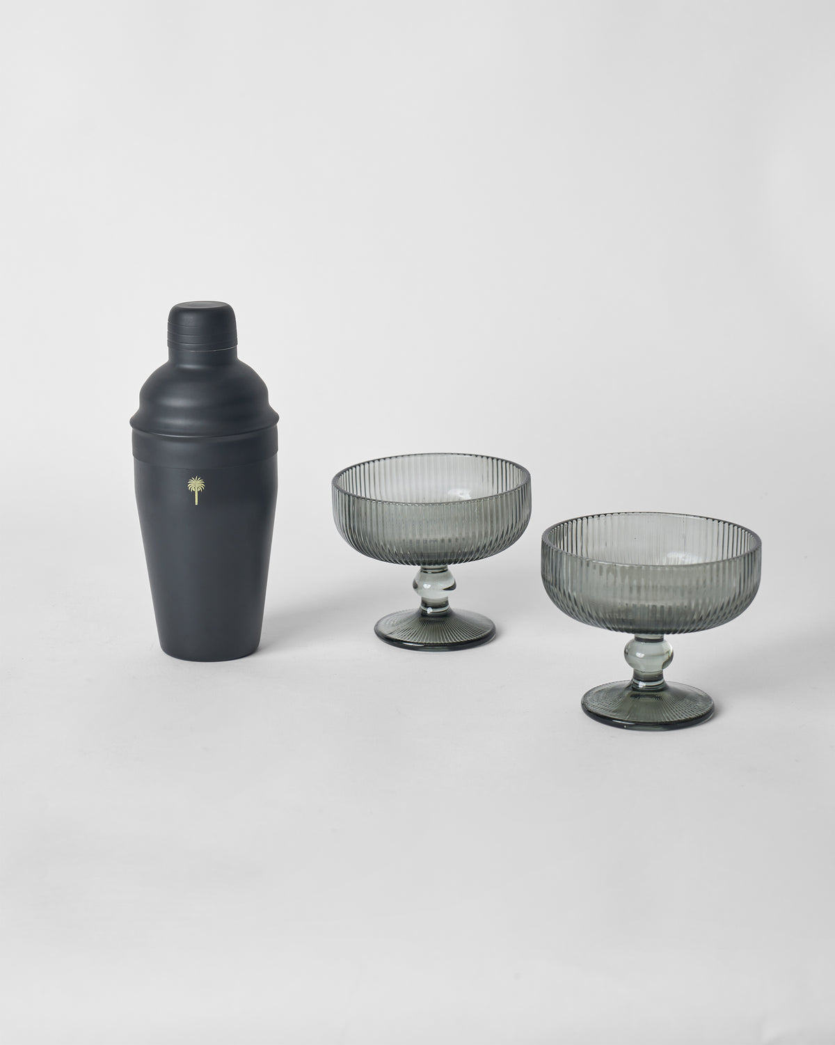 Black cocktail shaker with gold palm logo beside two smoked fluted coupe glasses.