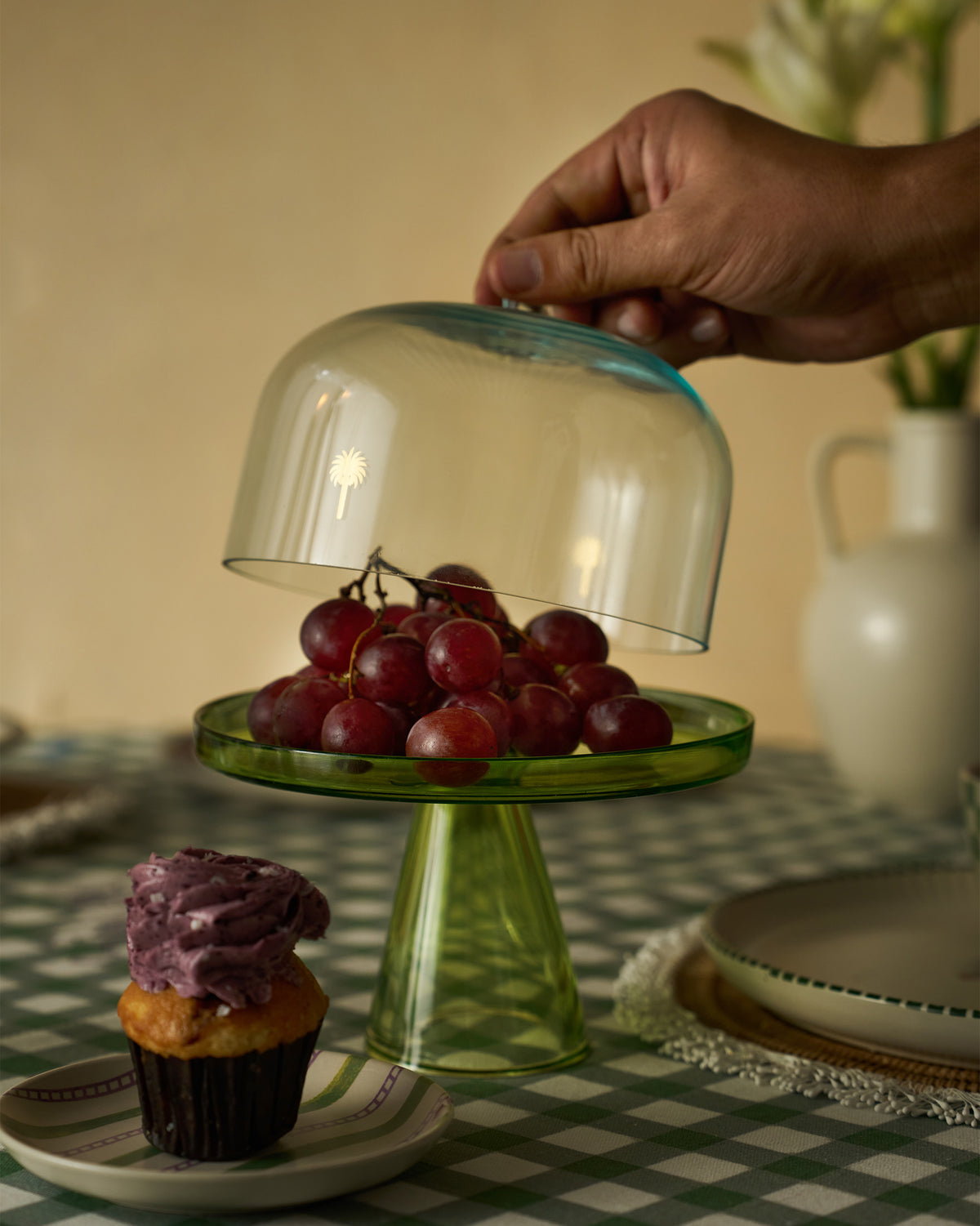 Hand lifting the glass dome on a Vatika cake stand filled with grapes.