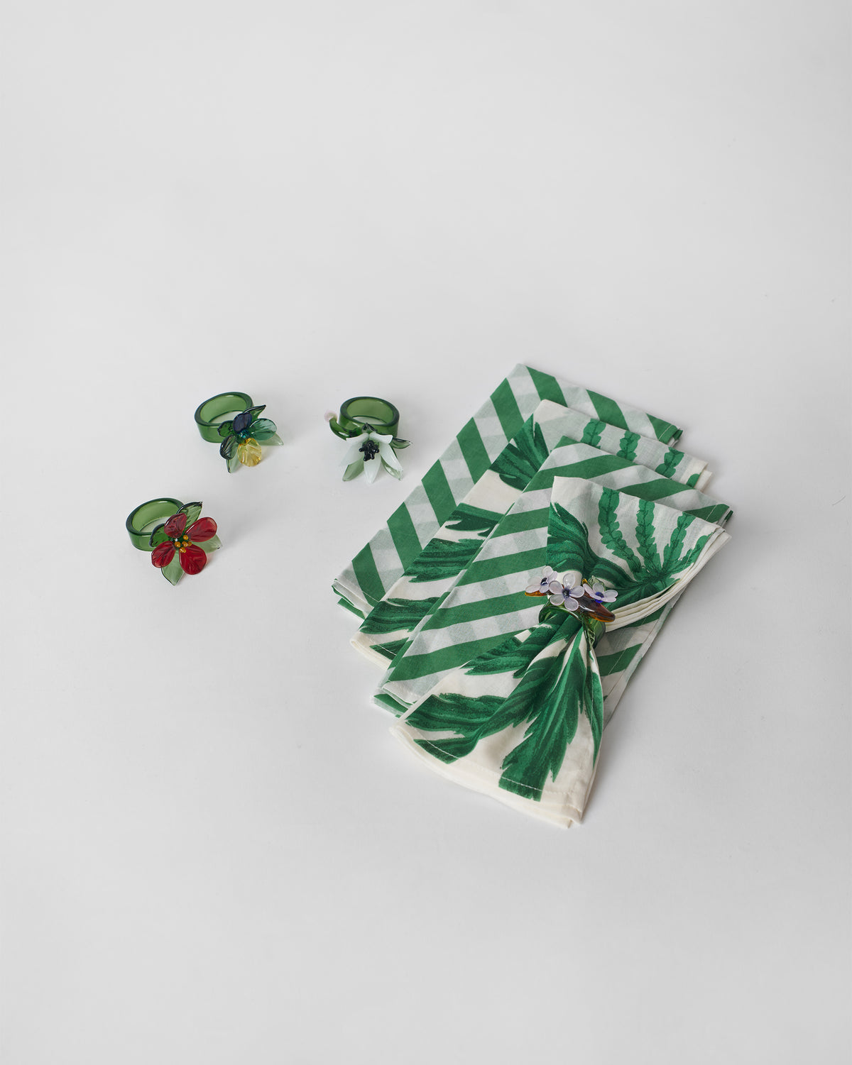 Folded Vanika Gift Set napkins in green stripe and palm prints beside flower napkin rings.
