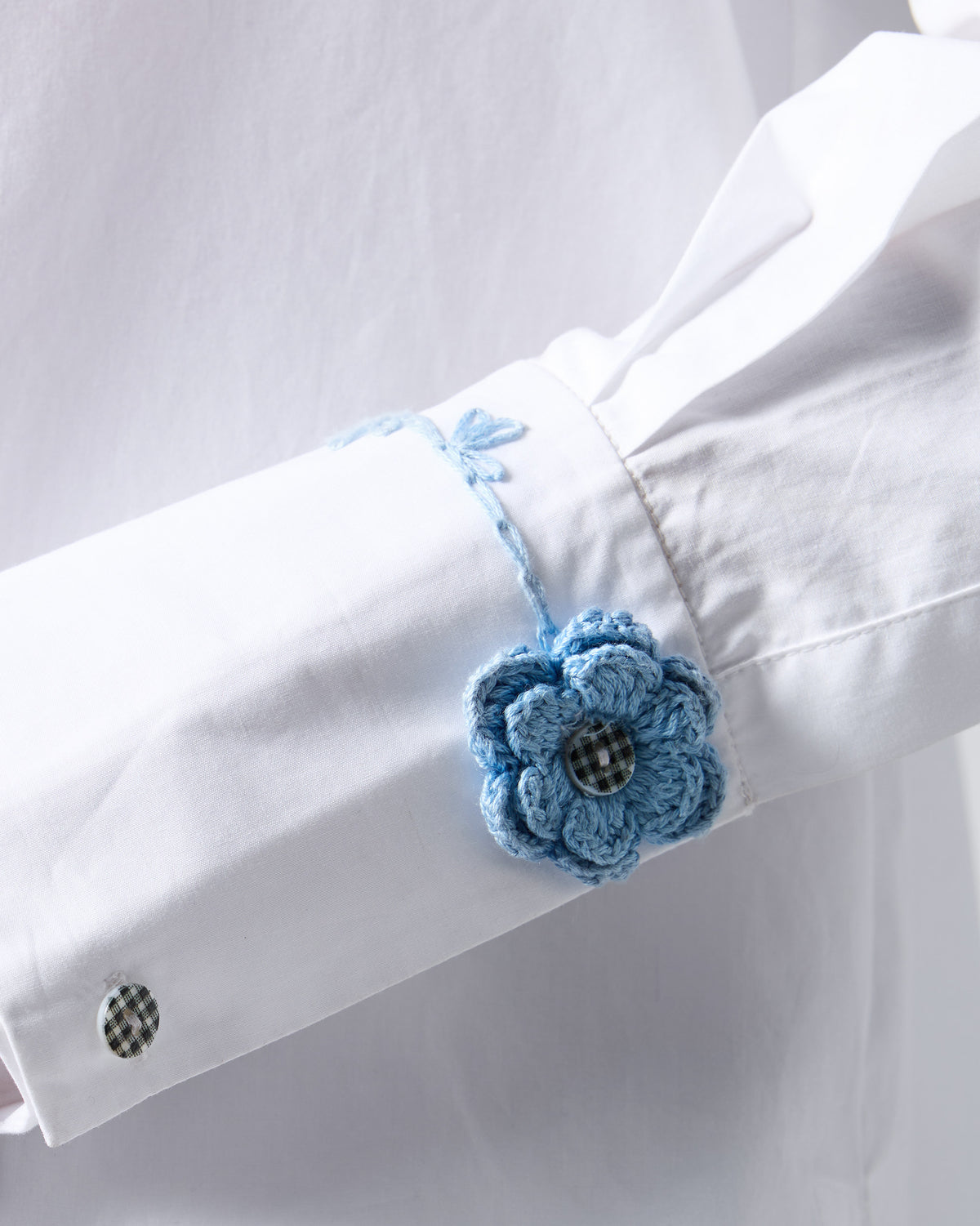 Close-up of a blue crochet flower on the cuff of a white shirt.
