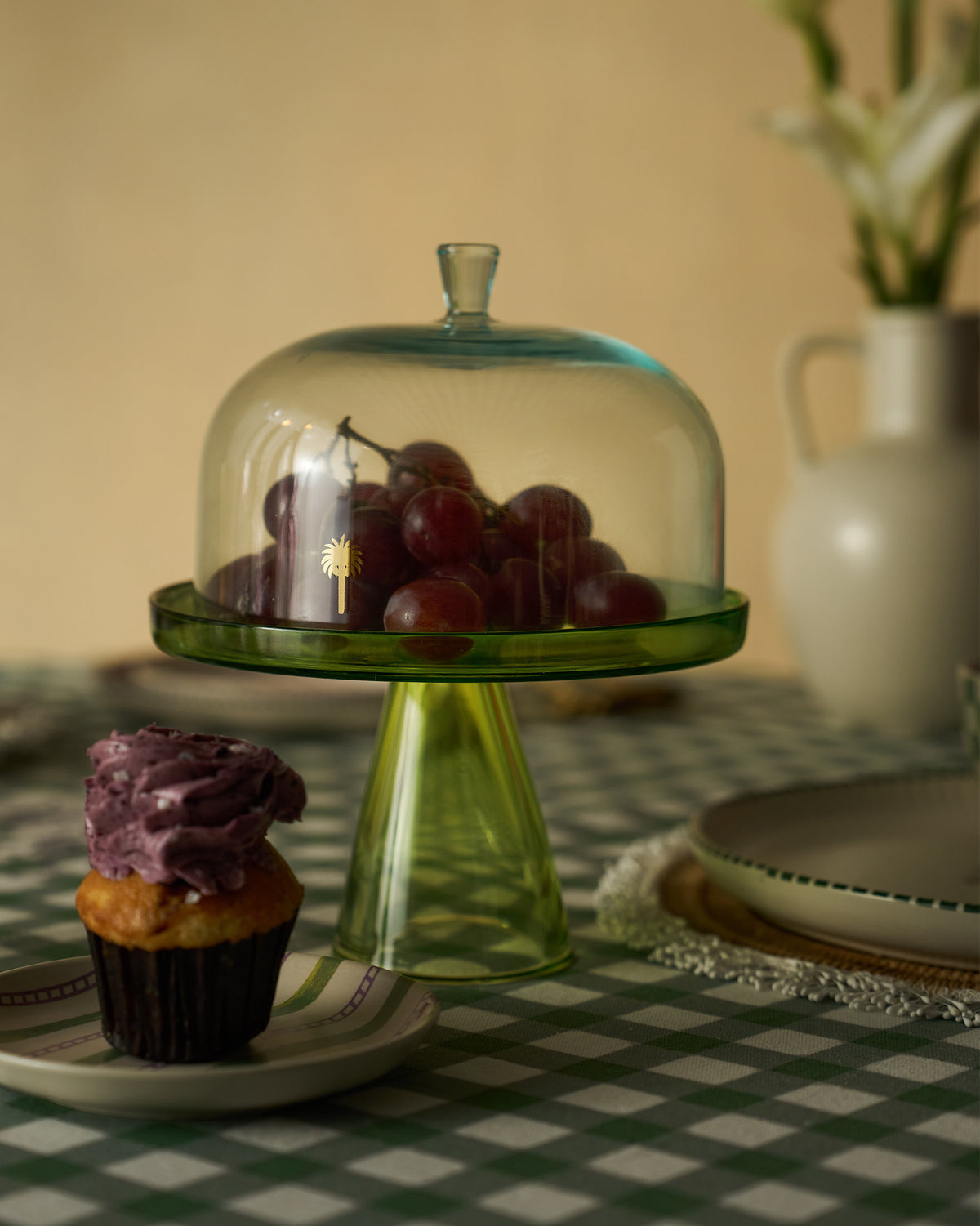 Green Vatika glass cake stand with dome and gold palm motif holding grapes.