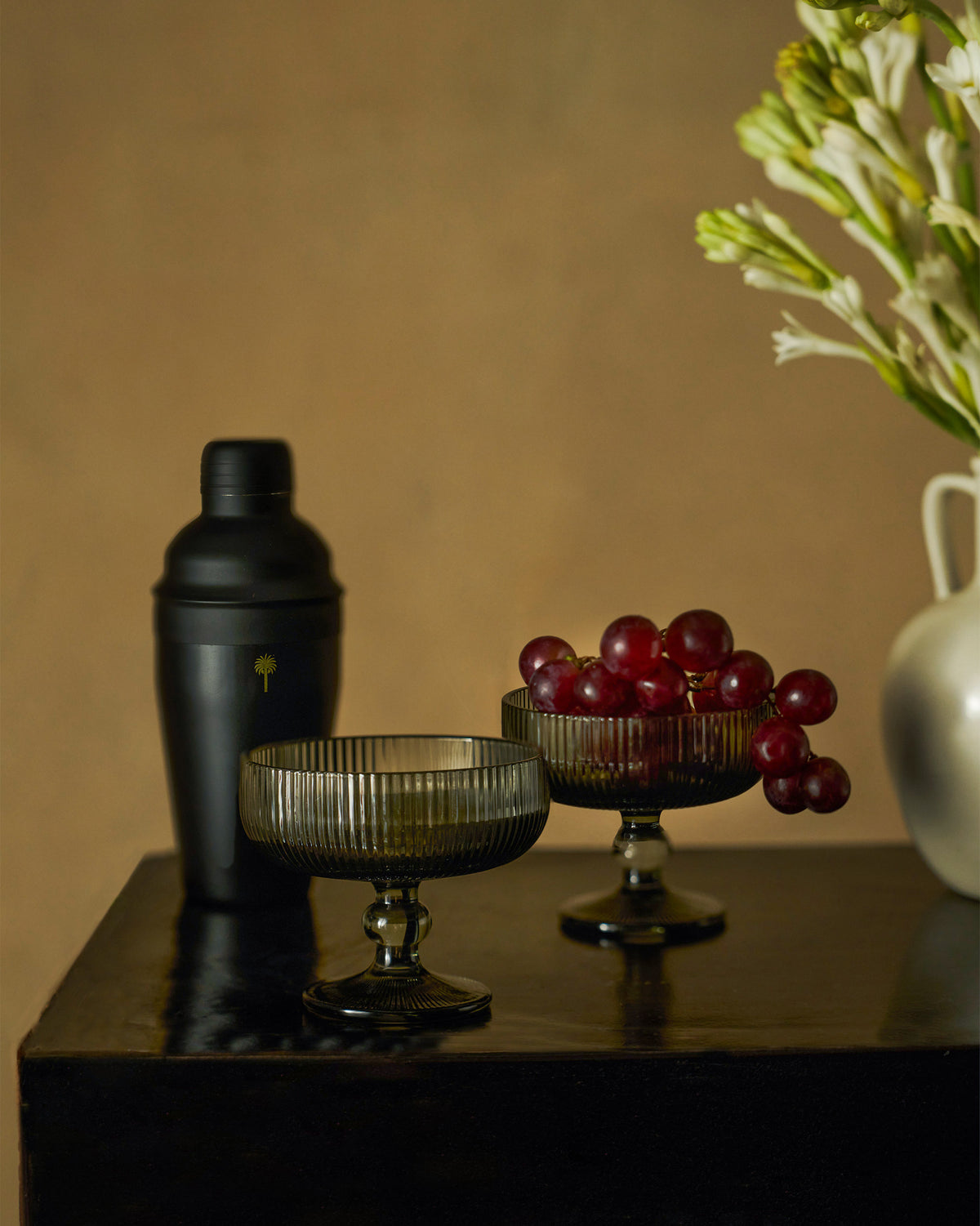 Avi smoked coupe glasses and black shaker styled with grapes on a dark table.