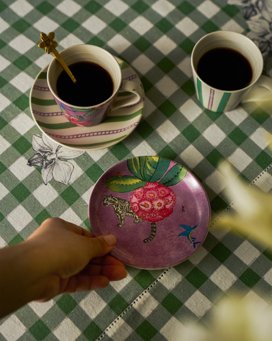 A hand holding a lavender Buransh plate on green gingham beside coffee