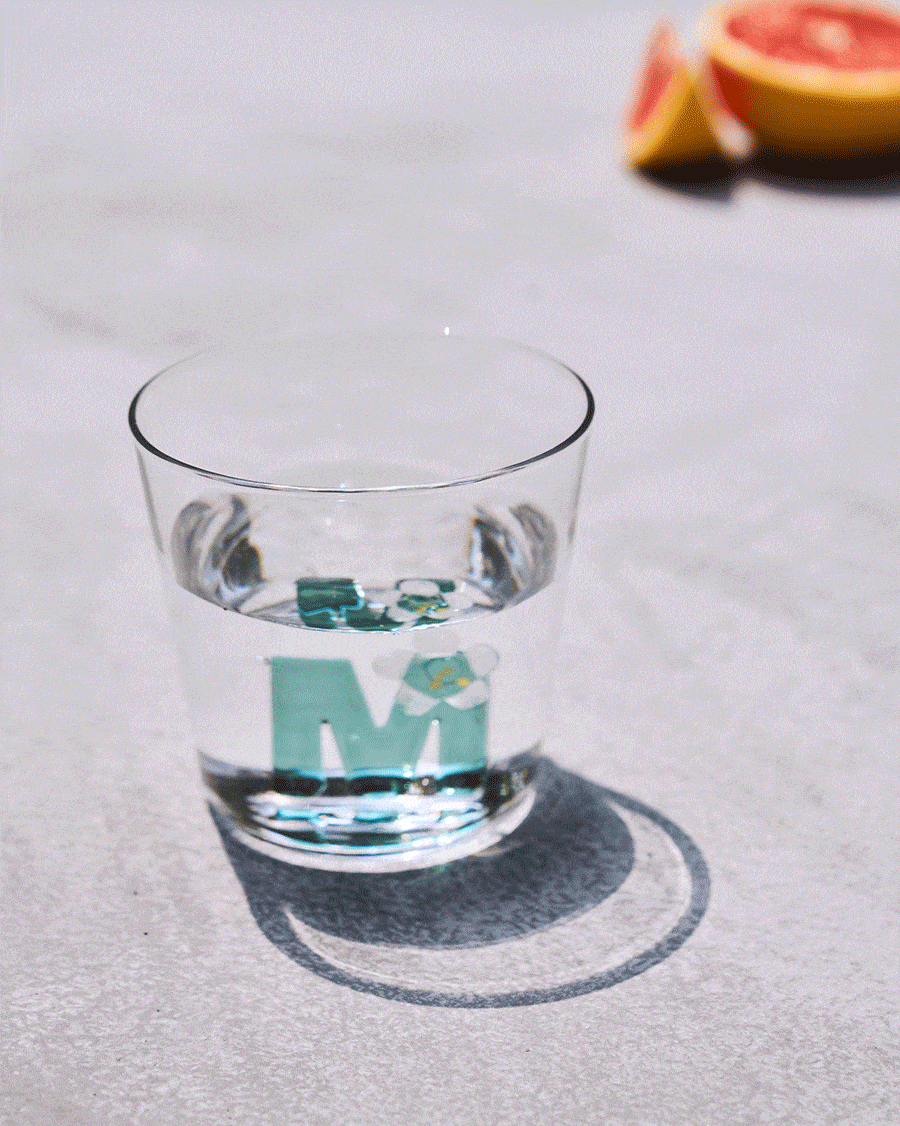 Clear Alphabet glass with a teal letter M and white flower, half filled with water.