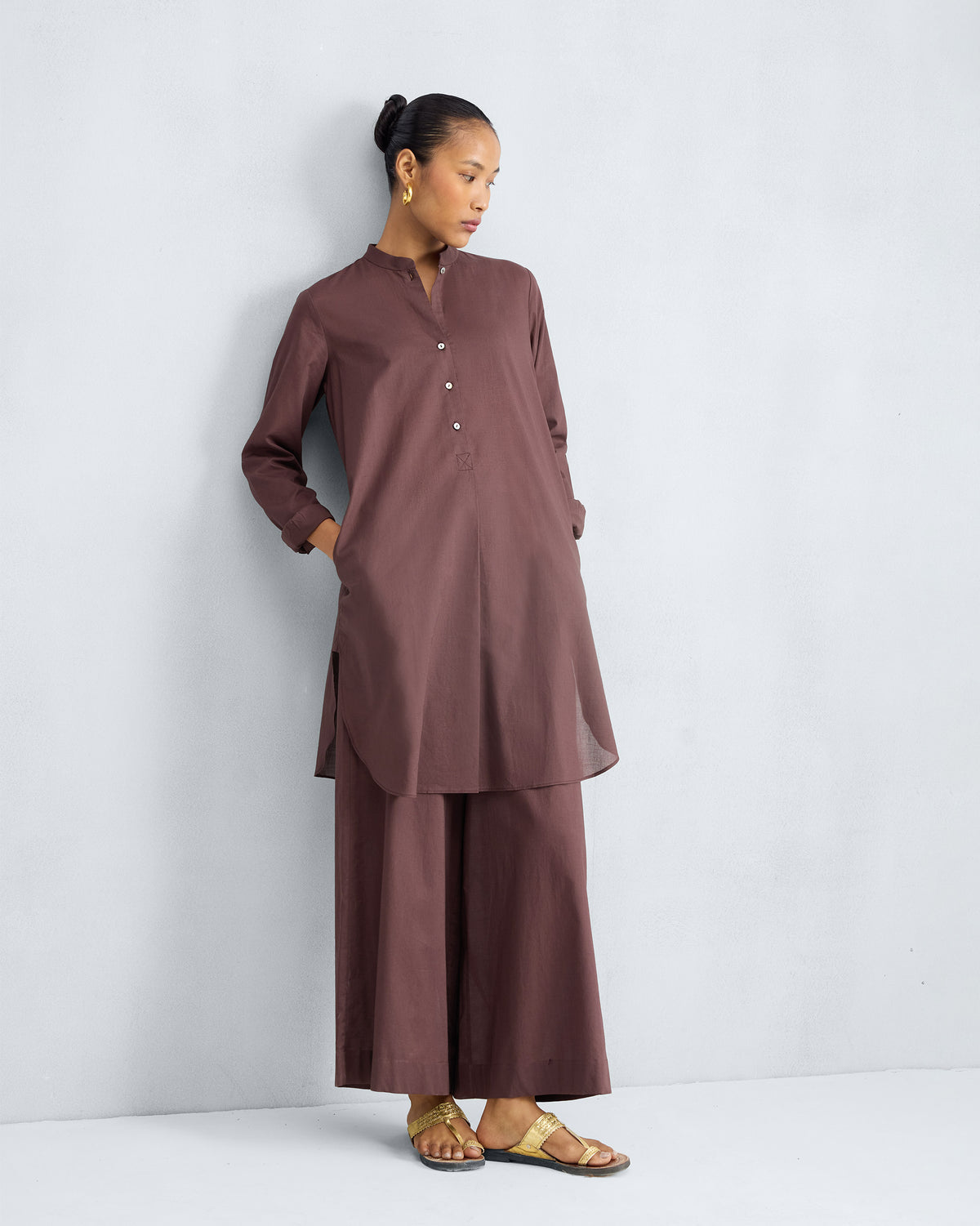 Person wearing a chocolate plum mid-length kurta set with hands in the pockets.