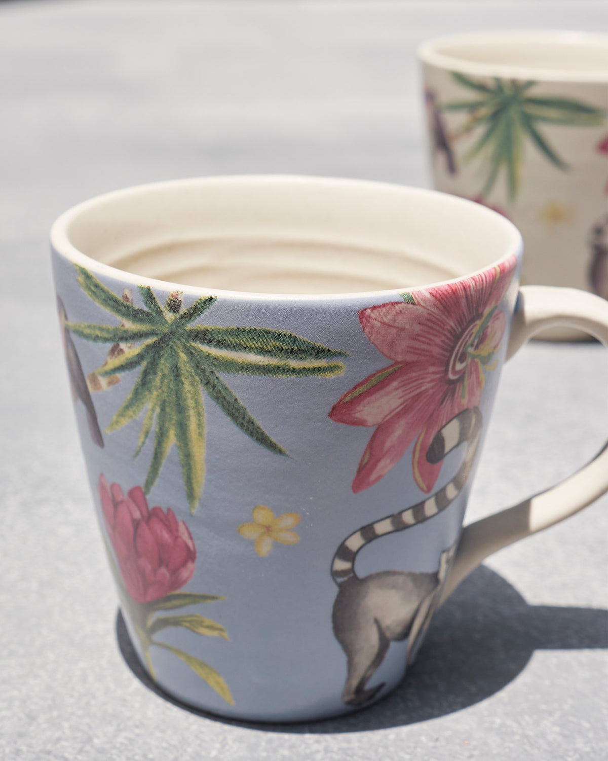 Blue mug with a ring-tailed lemur and pink flower beside a cream mug.