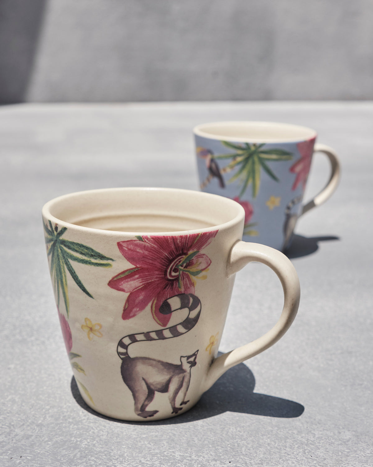 Cream mug with a lemur and pink flower beside a blue tropical mug.