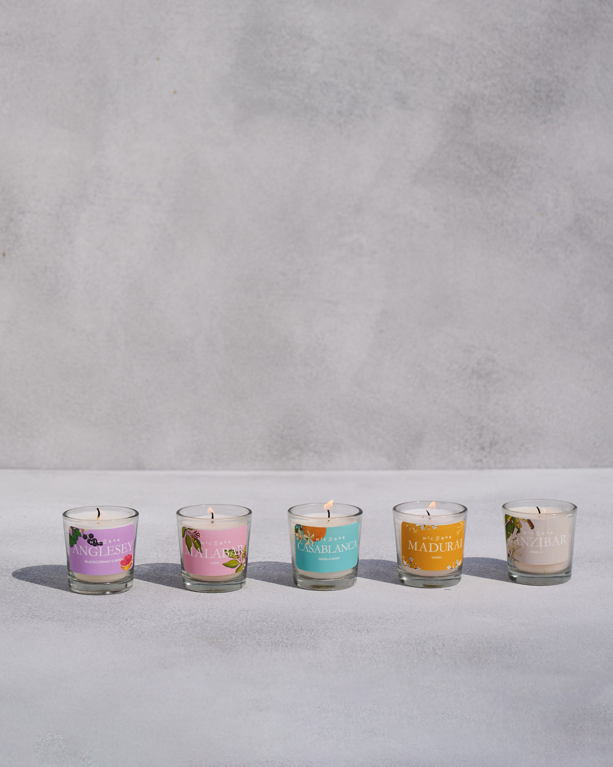 Five lit Nicobar jar candles in a row with coloured scent labels.