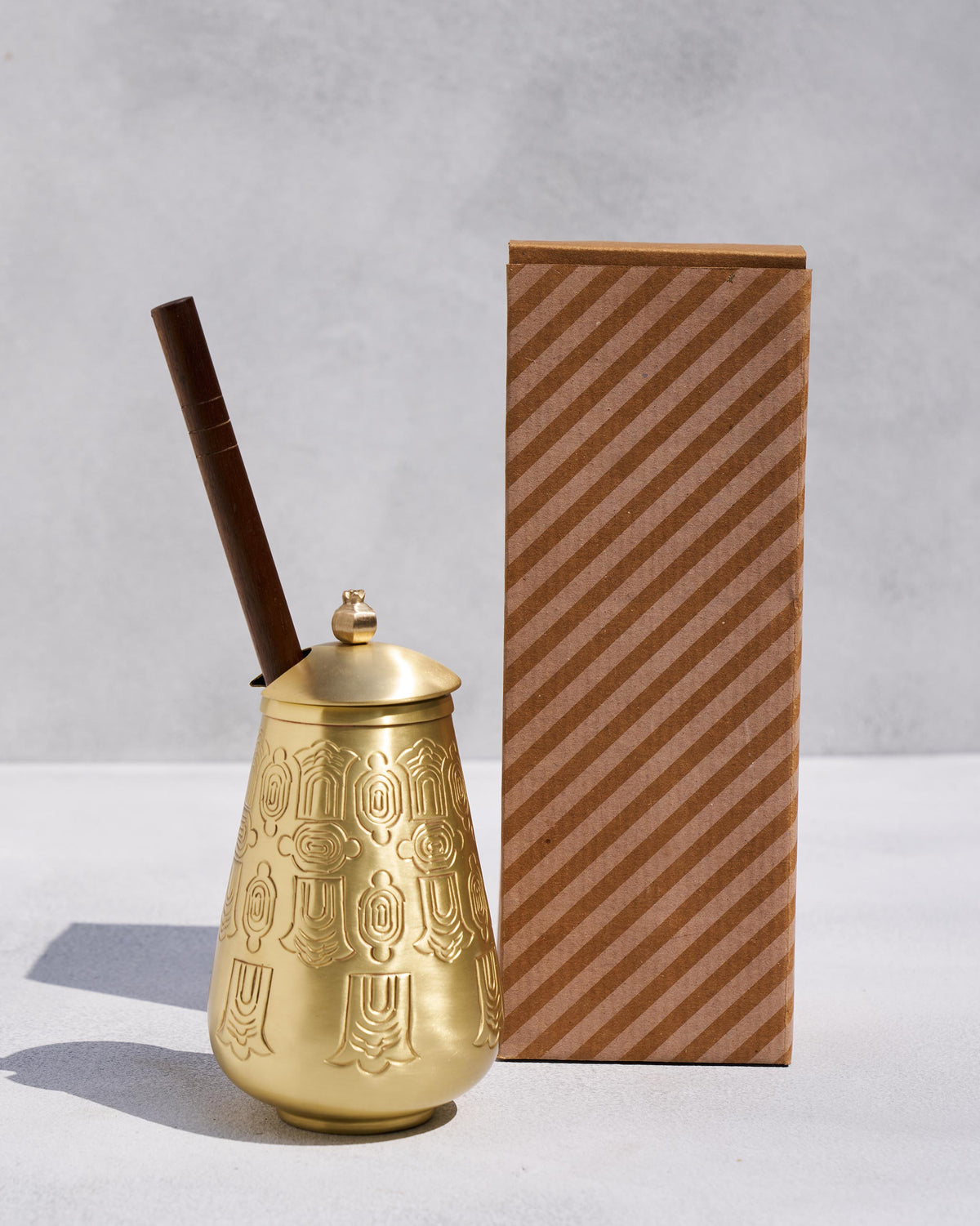 Gold embossed Dariya lota with wooden churner beside a striped brown gift box.