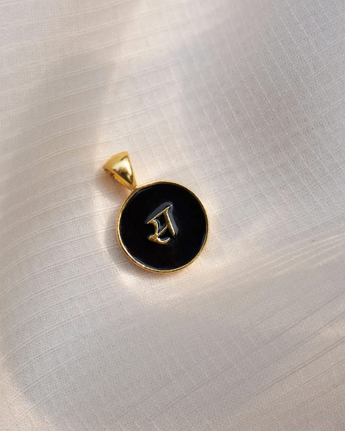 Close-up of a black enamel alphabet charm with a raised gold Devanagari स.