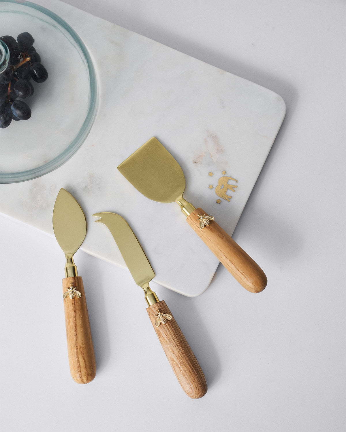 Garo cheese knives with bee charms resting on a white marble serving board.