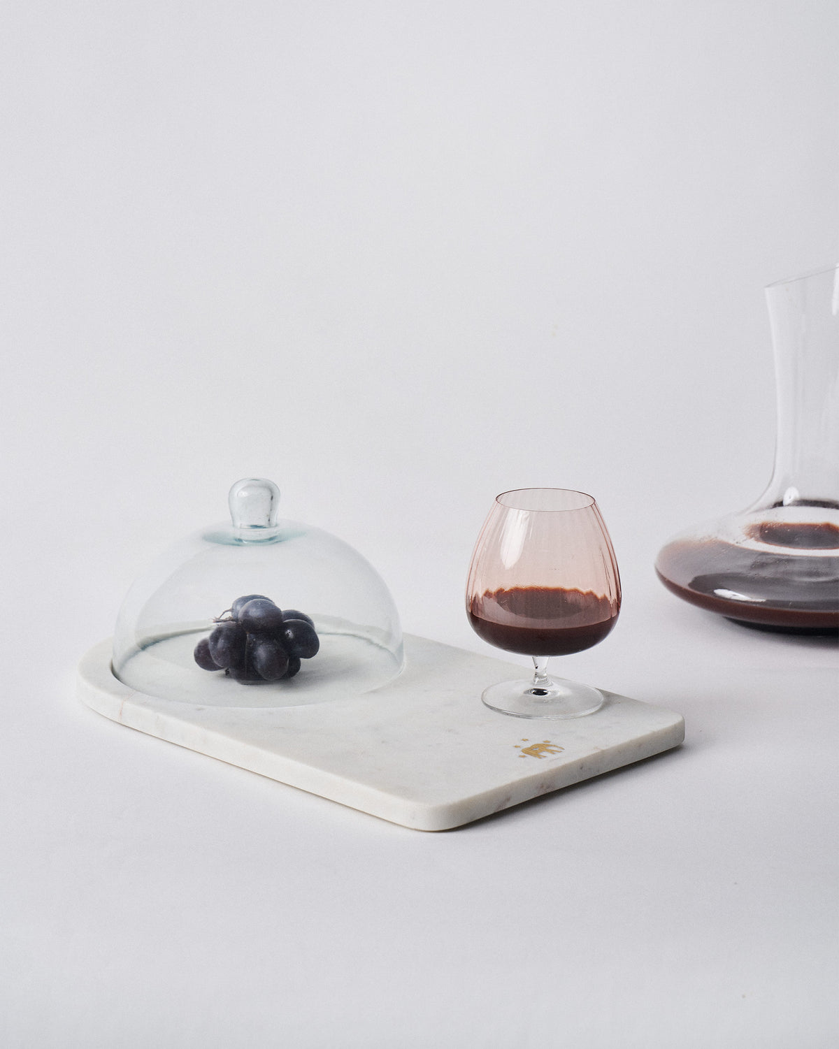 Ele cheese cloche covering grapes on marble beside a glass of wine.