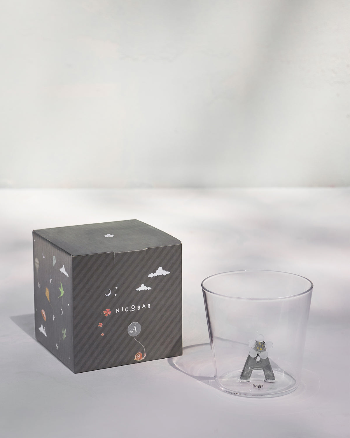 Clear alphabet glass with a 3D letter A beside a charcoal Nicobar gift box.