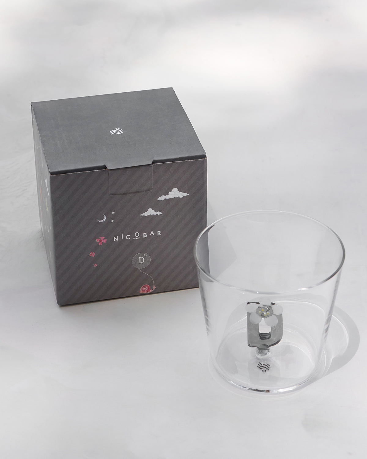 Clear Alphabet glass with a 3D letter D beside a charcoal Nicobar gift box.