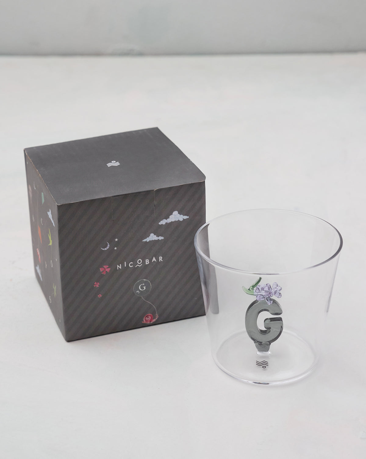 Clear Alphabet glass with a 3D letter G beside a charcoal Nicobar gift box.