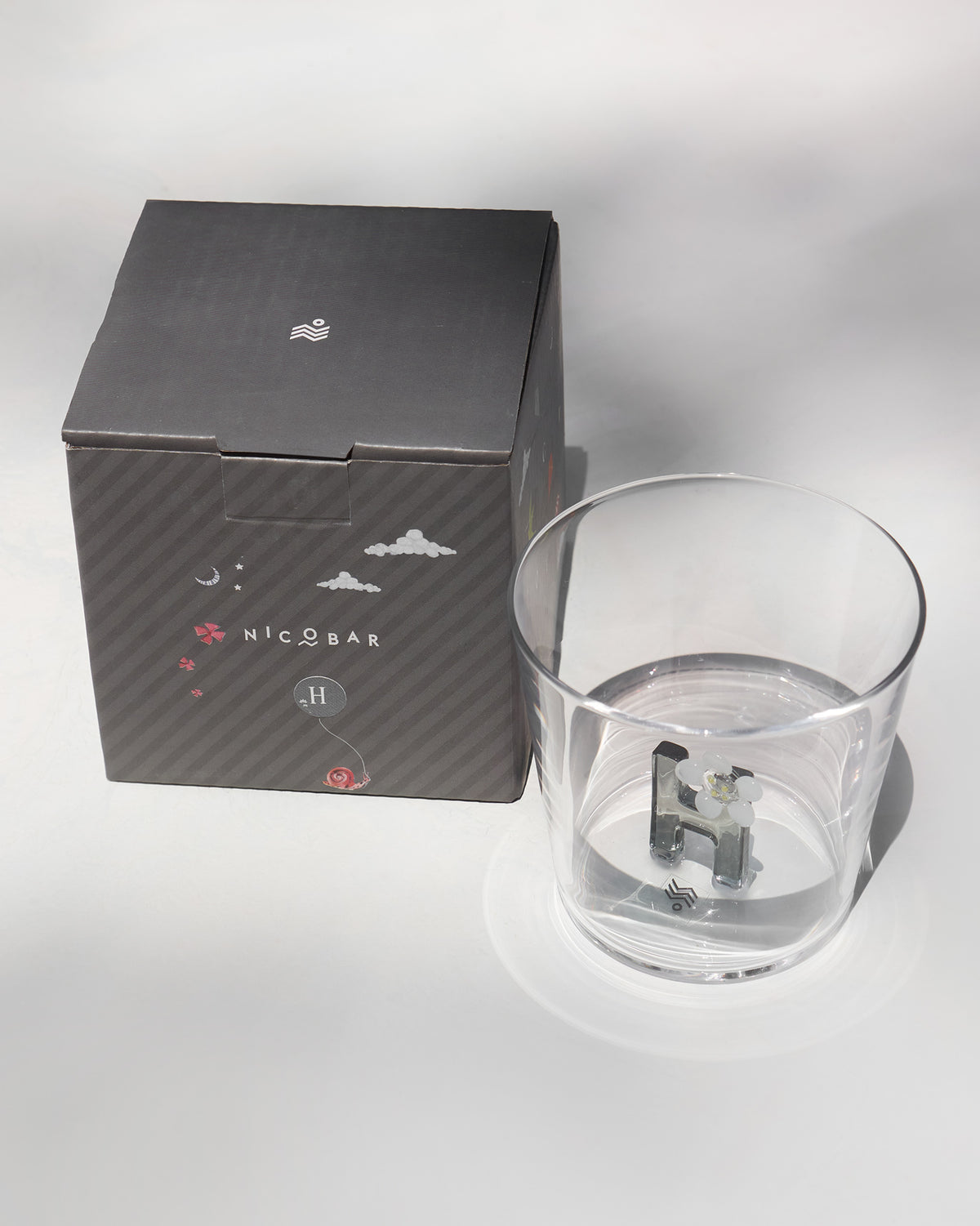Clear Alphabet glass with a 3D letter H beside a charcoal Nicobar gift box.