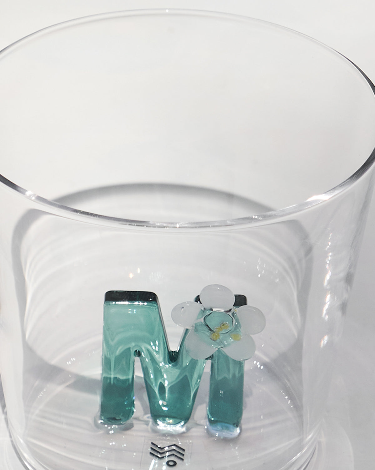 Close-up of the teal letter M with a white flower inside a clear Alphabet glass.