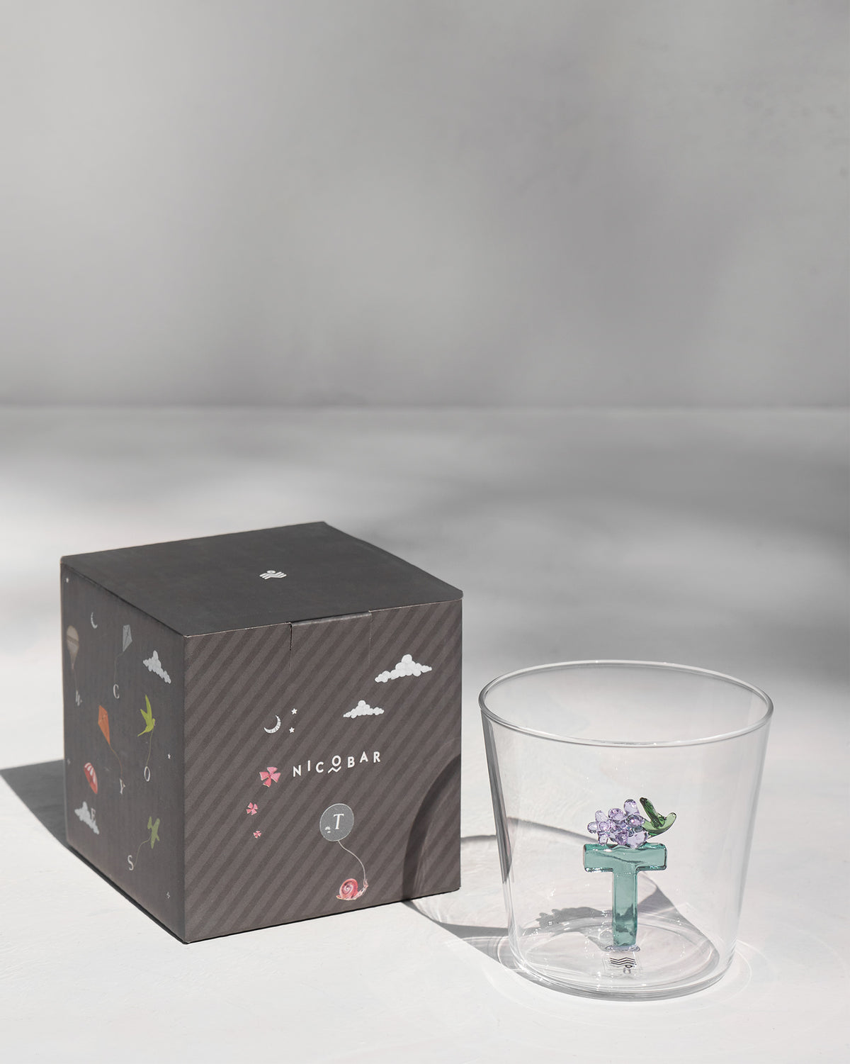 Alphabet glass with a teal letter T beside its charcoal Nicobar gift box.