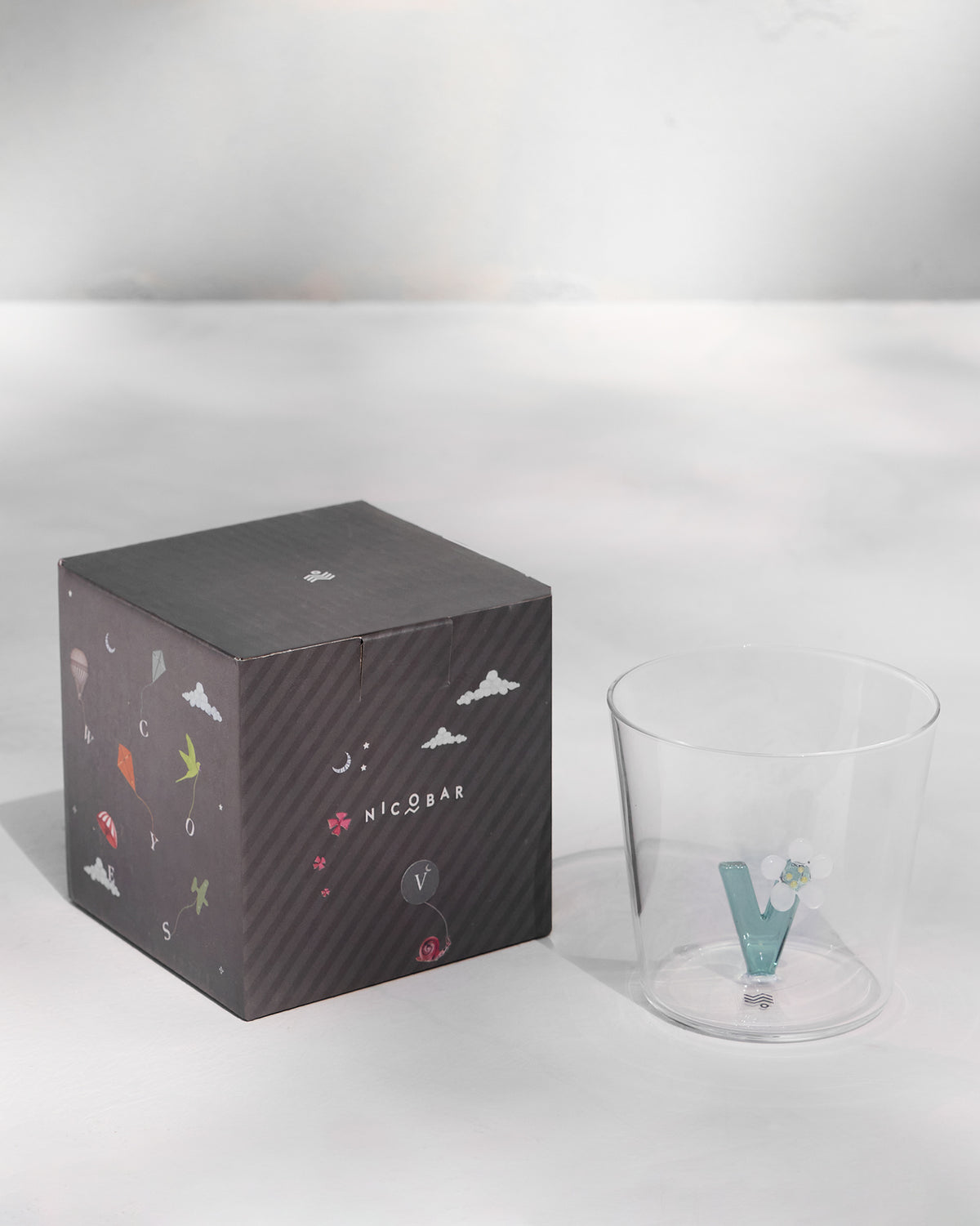 Alphabet glass with a teal letter V beside its charcoal Nicobar gift box.
