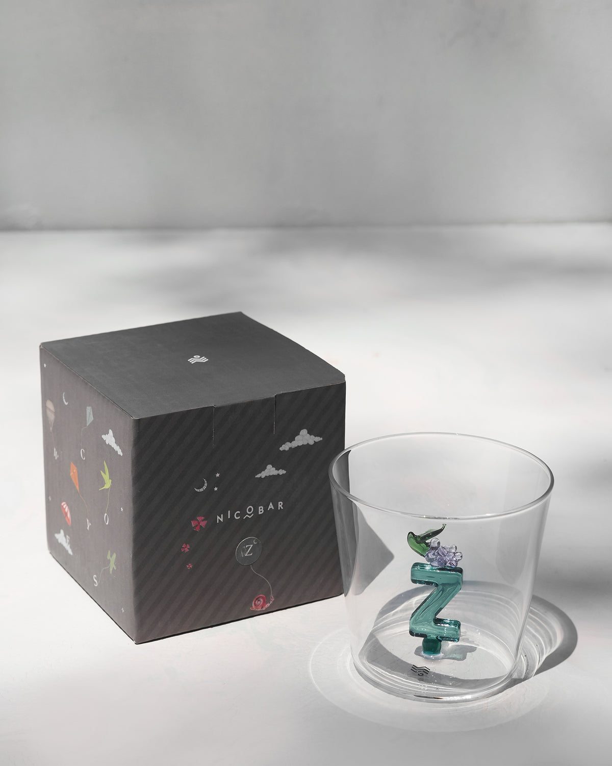 Alphabet glass with a teal letter Z beside its charcoal Nicobar gift box.
