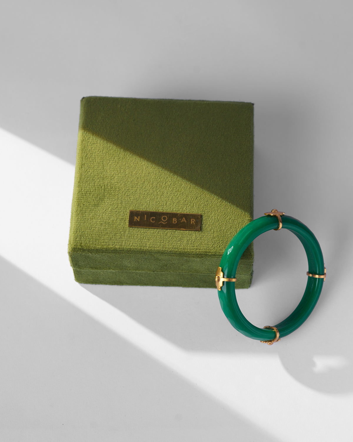 Green glass bangle with gold accents leaning on an olive velvet Nicobar box.