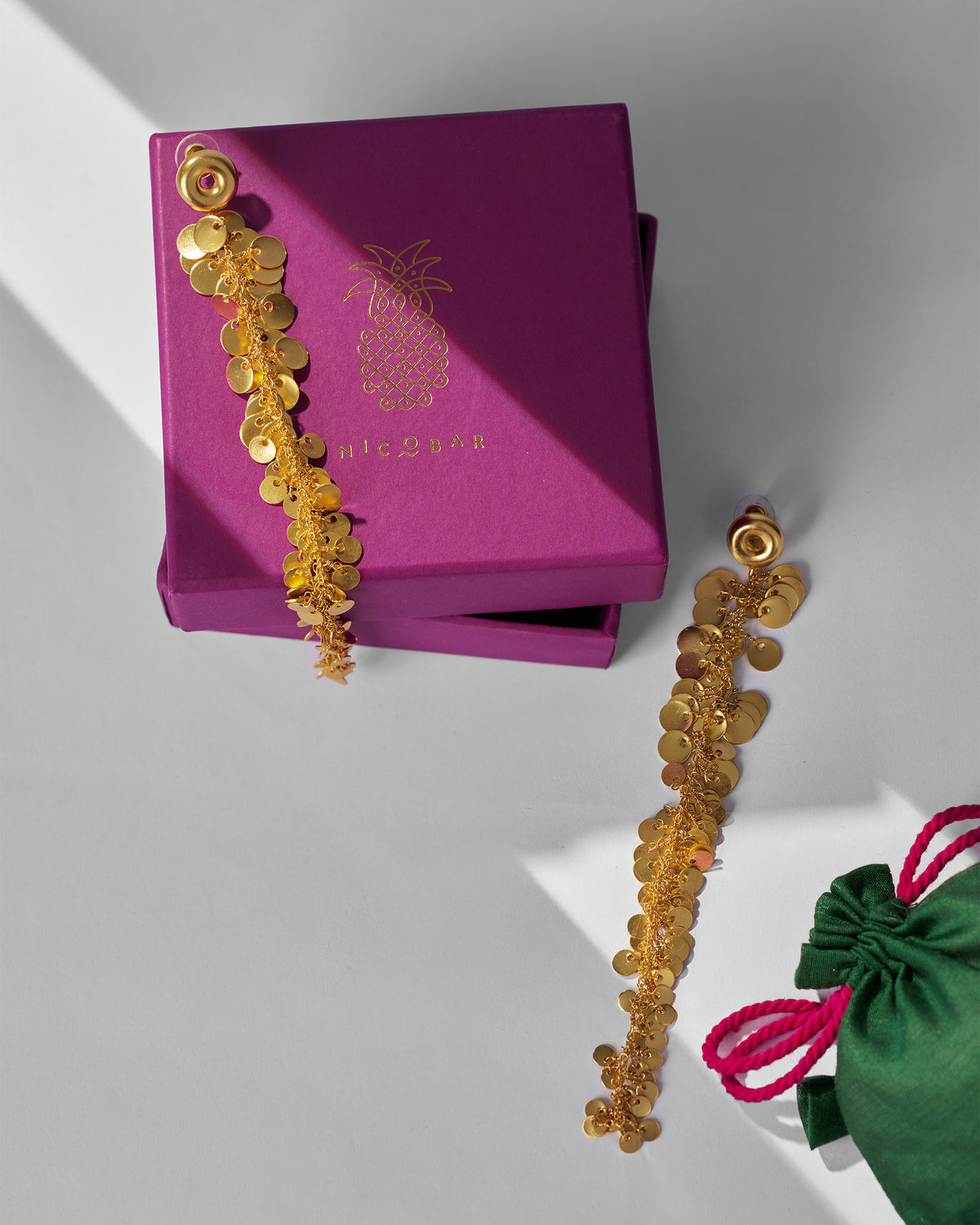 Pair of gold Tassel Shoulder Dusters draped beside a magenta Nicobar gift box.