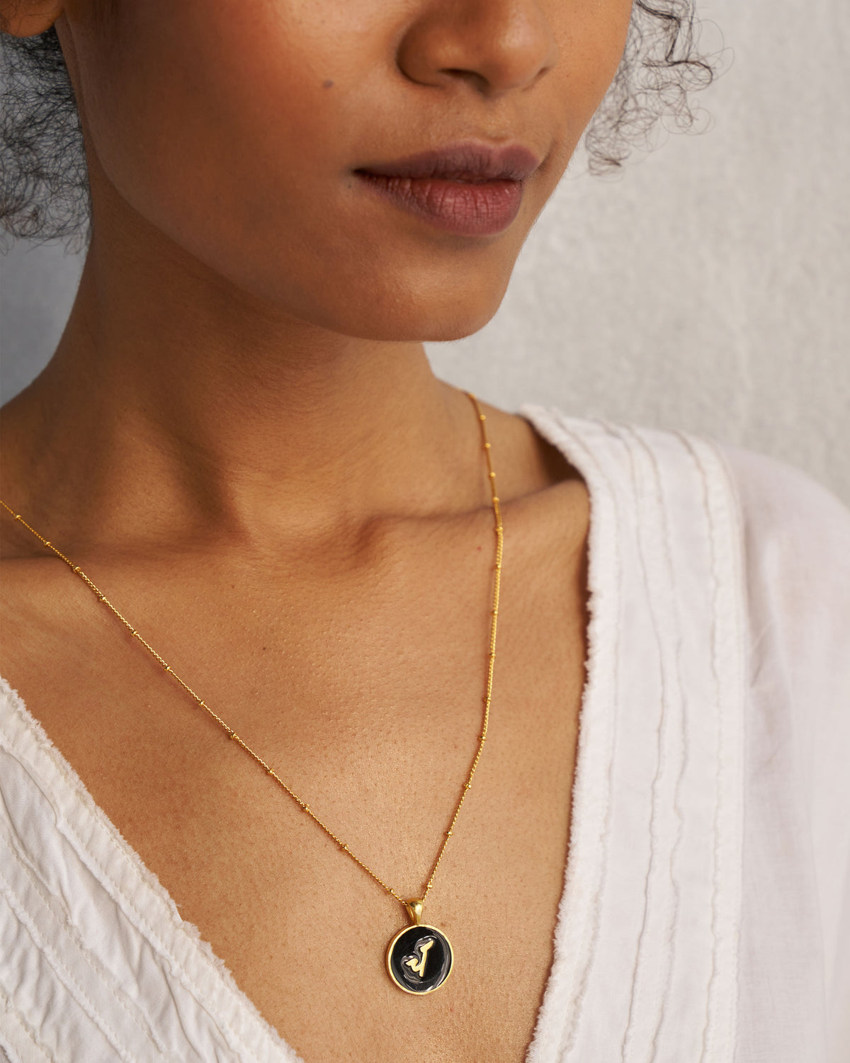 Gold alphabet charm with a black enamel disc worn on a beaded chain.