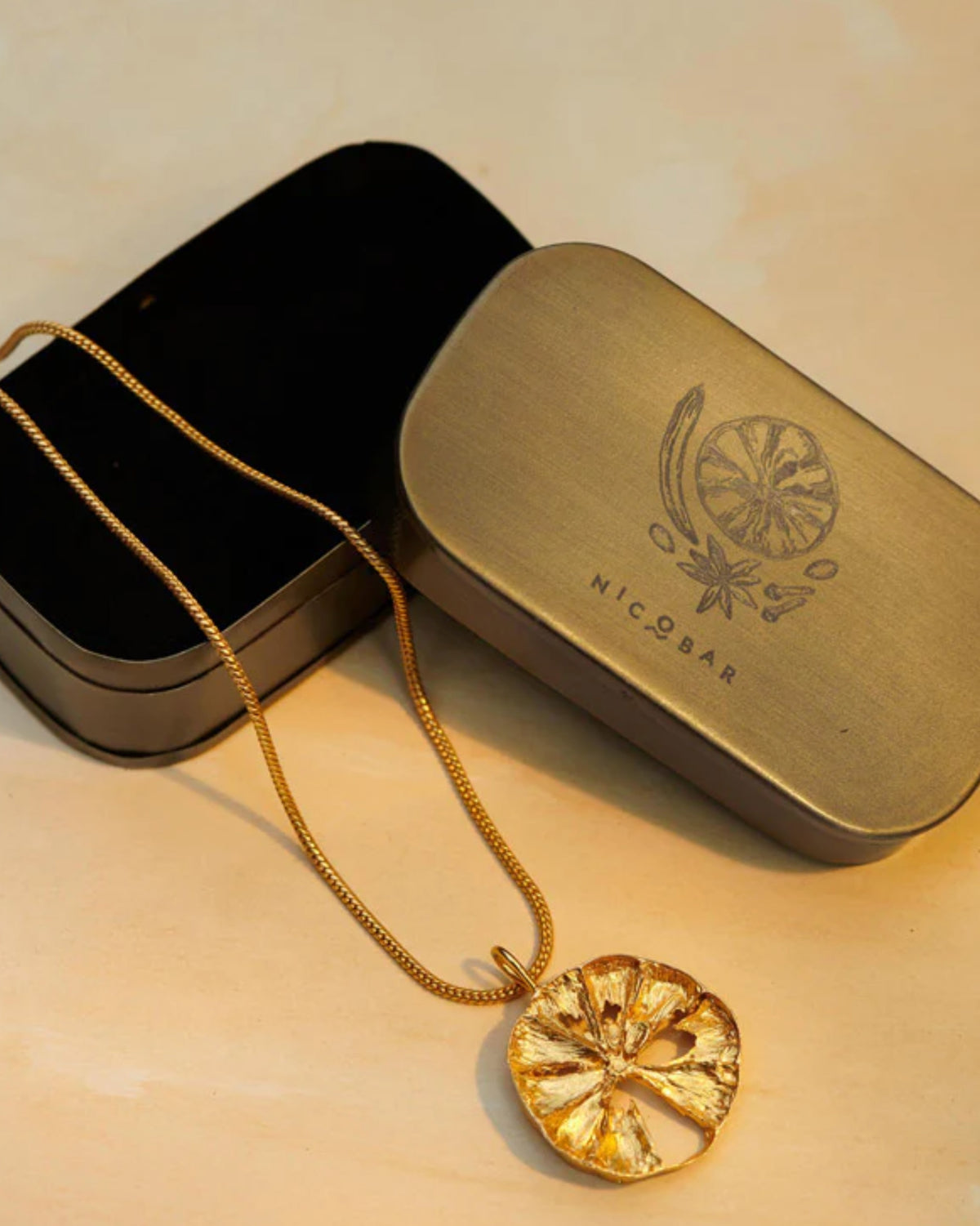 Gold lemon slice charm beside its Nicobar spice-illustrated gift tin.