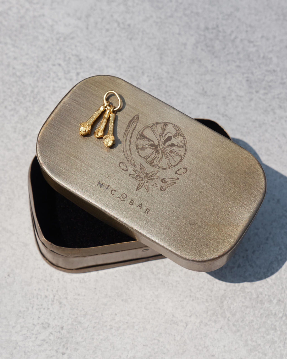 Three gold clove charms on a Nicobar spice-illustrated gift tin.