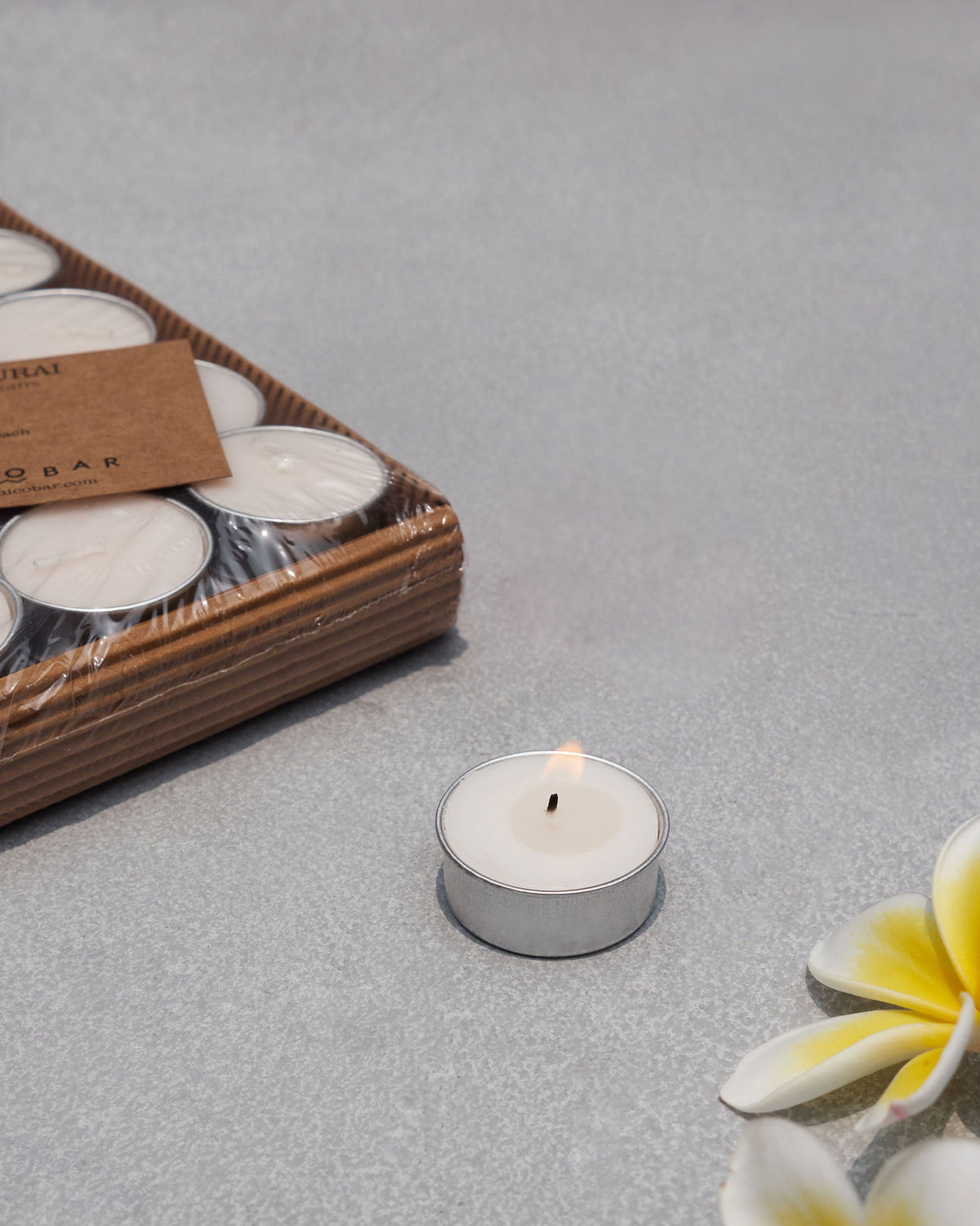 Madurai tealight pack with one lit candle and white blossoms nearby.