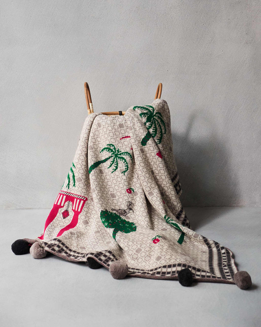 The Mirage throw draped over a cane chair, pompoms at the corners