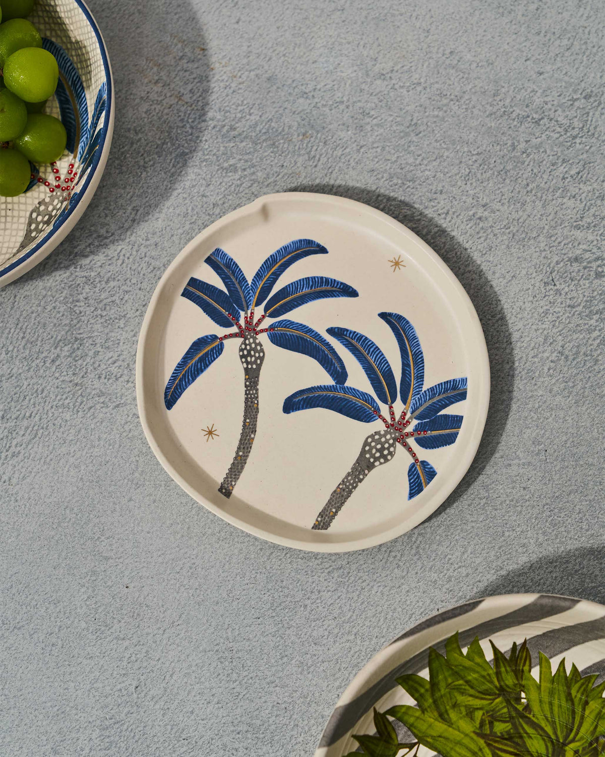 Dancing Palm Dessert Plate with blue palm trees, styled beside a bowl of grapes.