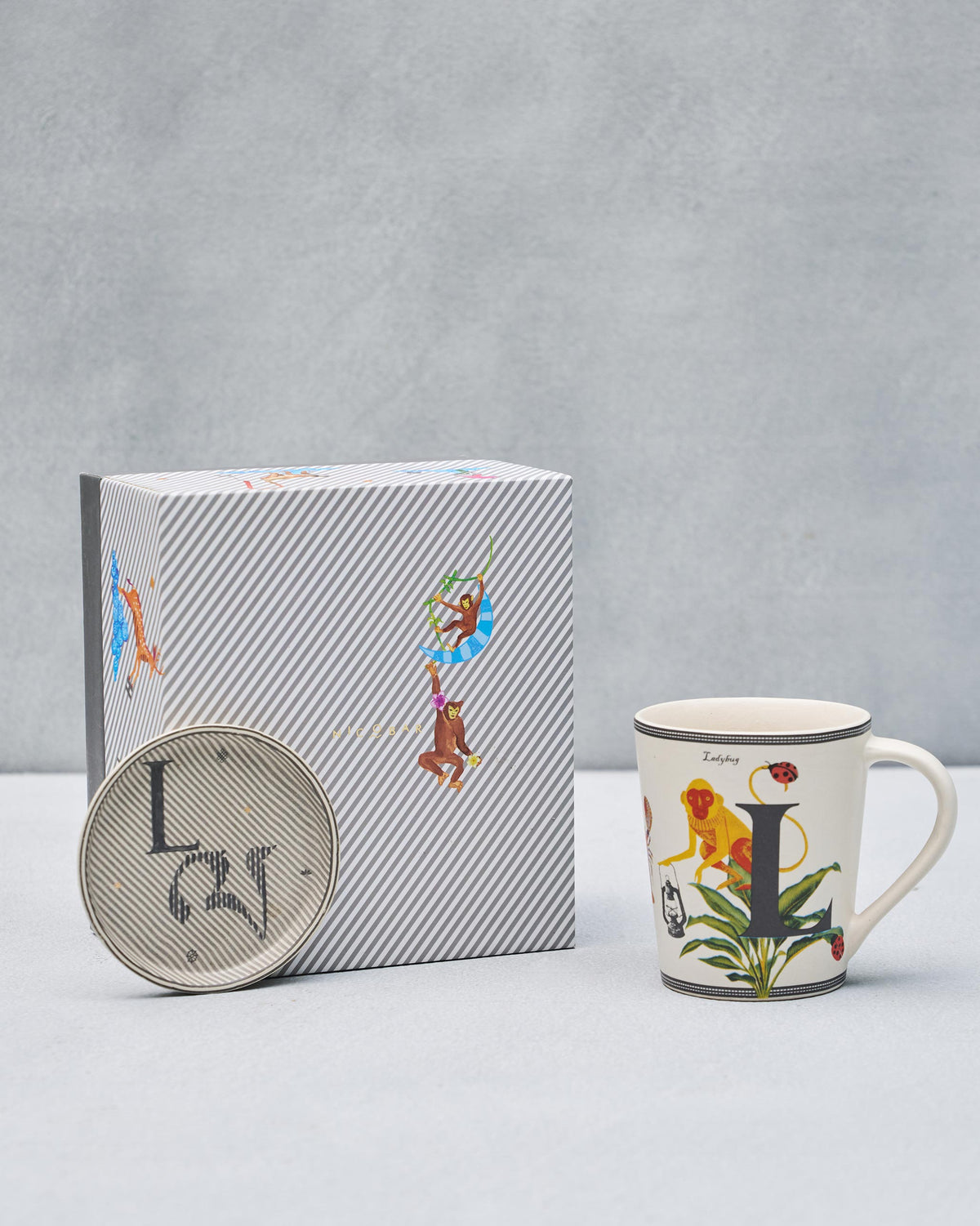 L alphabet mug with monkey and ladybug beside matching coaster and gift box.